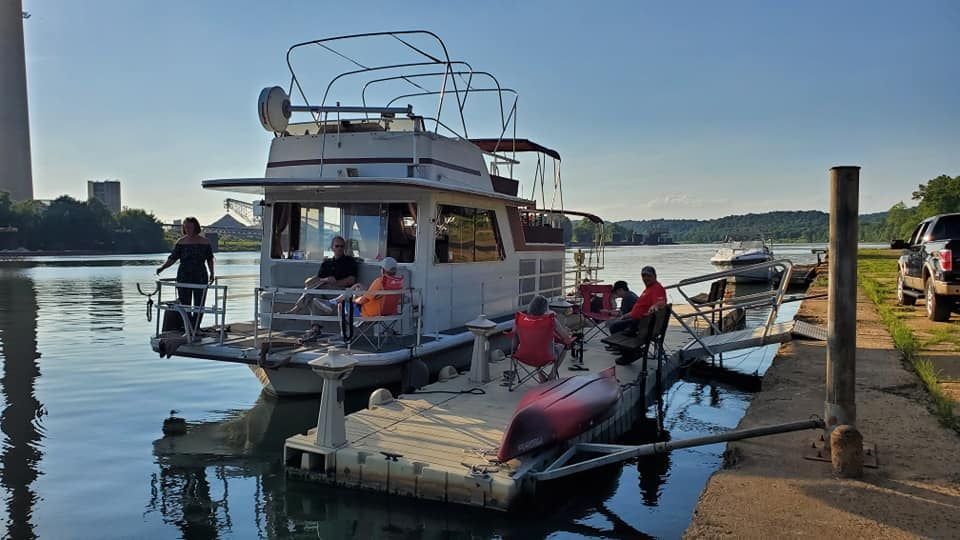 A houseboat docked at a pier with people sitting on the deck and the pier, next to a parked truck under a blue sky.