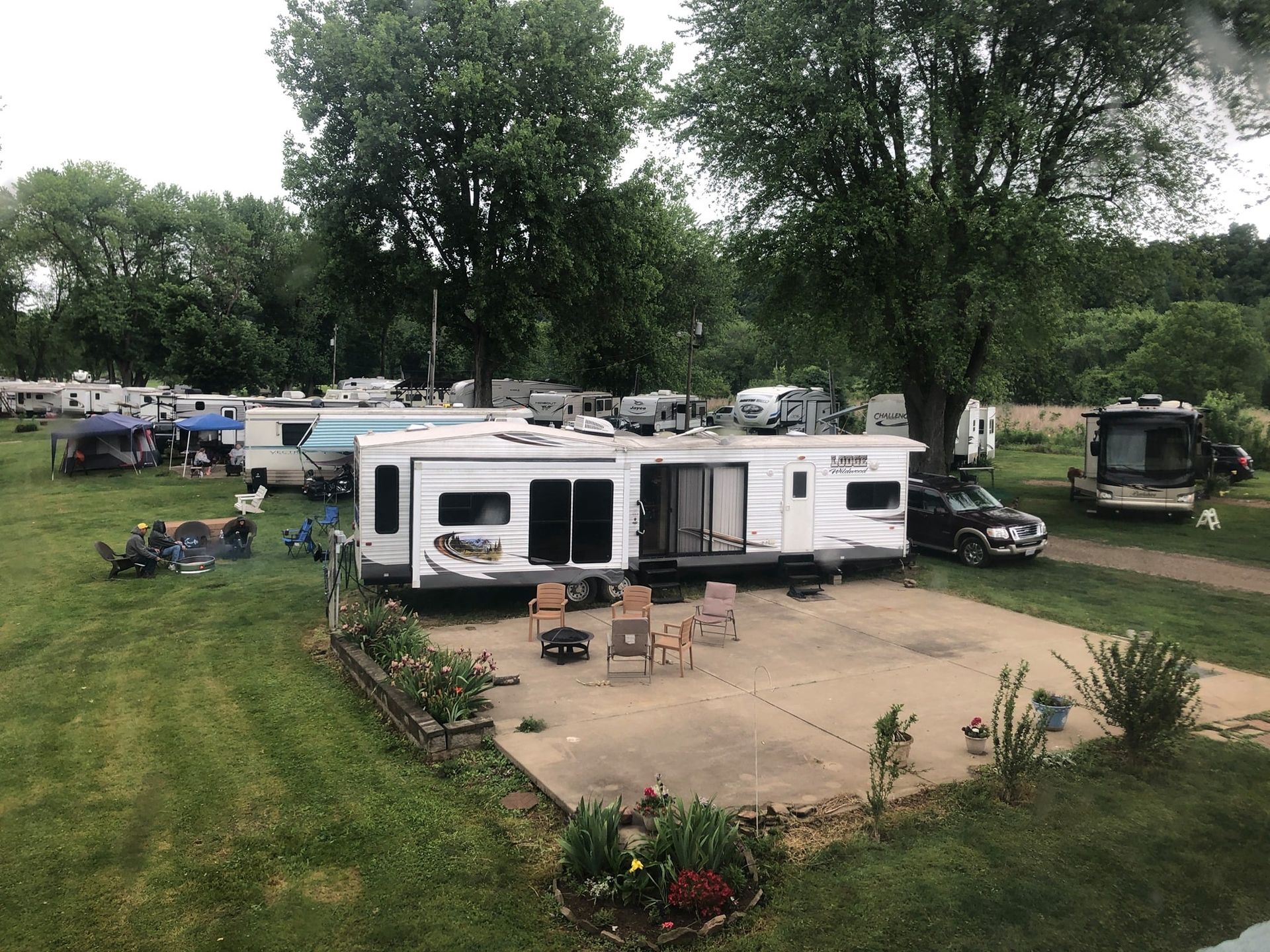 Camper trailer parked on a grassy campsite with chairs on a concrete pad, surrounded by trees and RVs