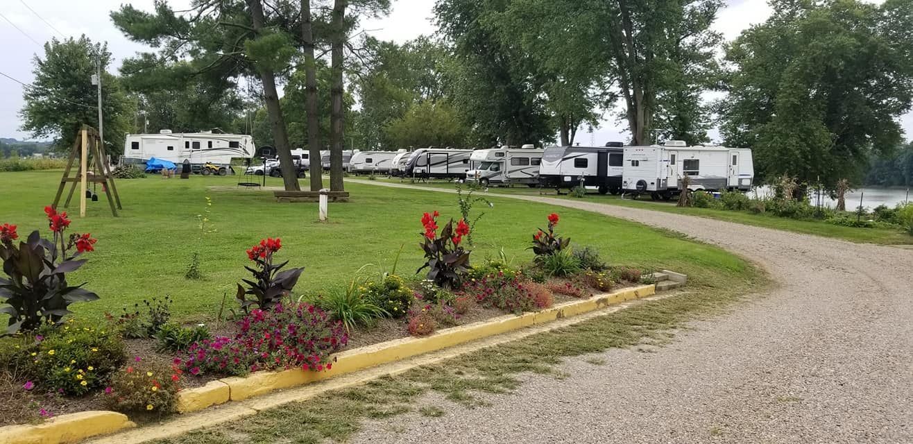 A gravel driveway lined with colorful flowers leads to a row of RVs parked on a grassy lot with trees near a lake.
