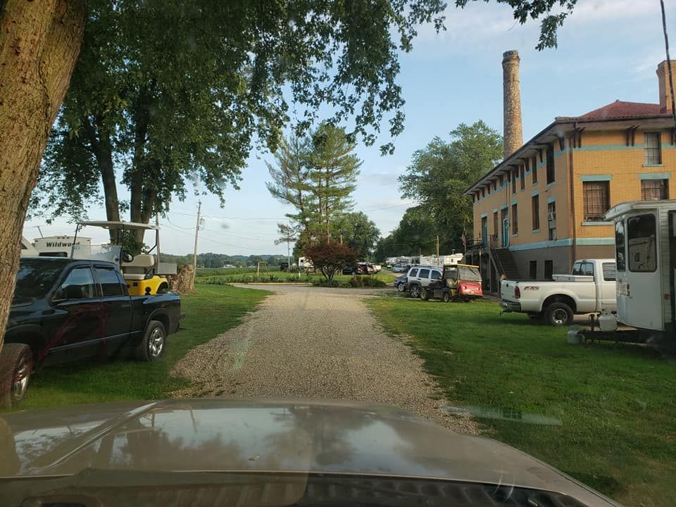 A gravel driveway leads toward trees and a yellow building with a tall brick chimney, flanked by parked trucks and trailers.