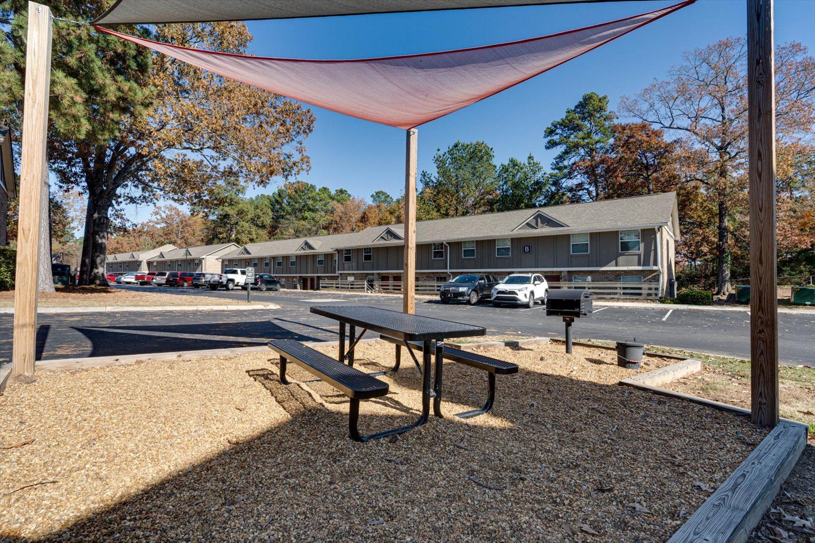 Outdoor picnic table and benches under shade sails in a gravel courtyard beside apartment buildings and parked cars