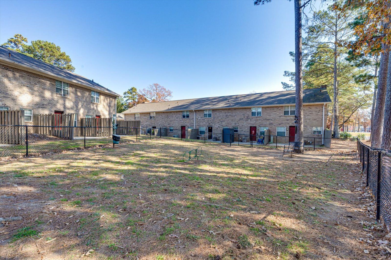 Backyard between brick buildings with fenced yard, trees, and bare ground in daylight