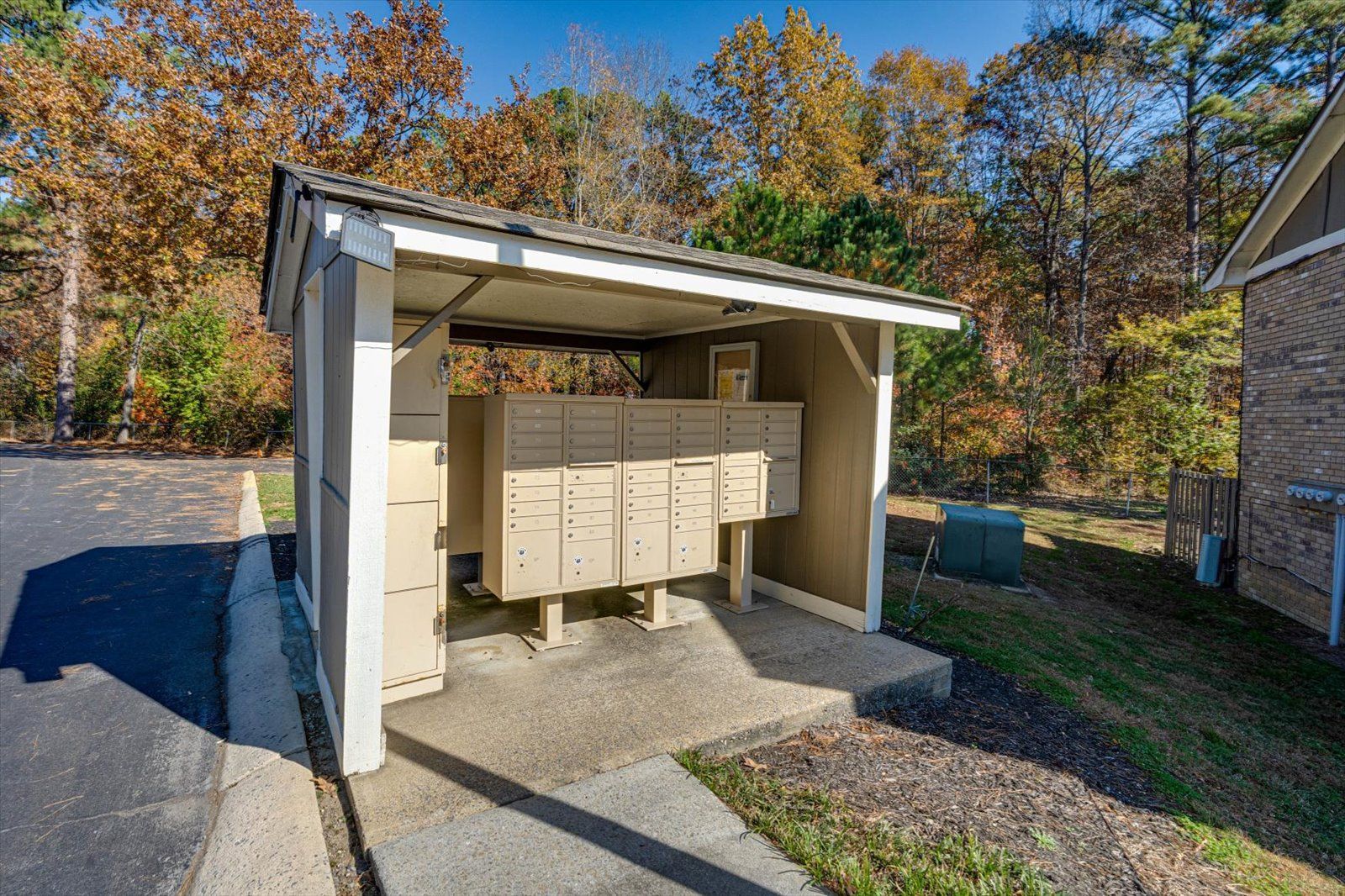 Outdoor beige mailbox cluster under a shelter beside a driveway, with autumn trees in the background