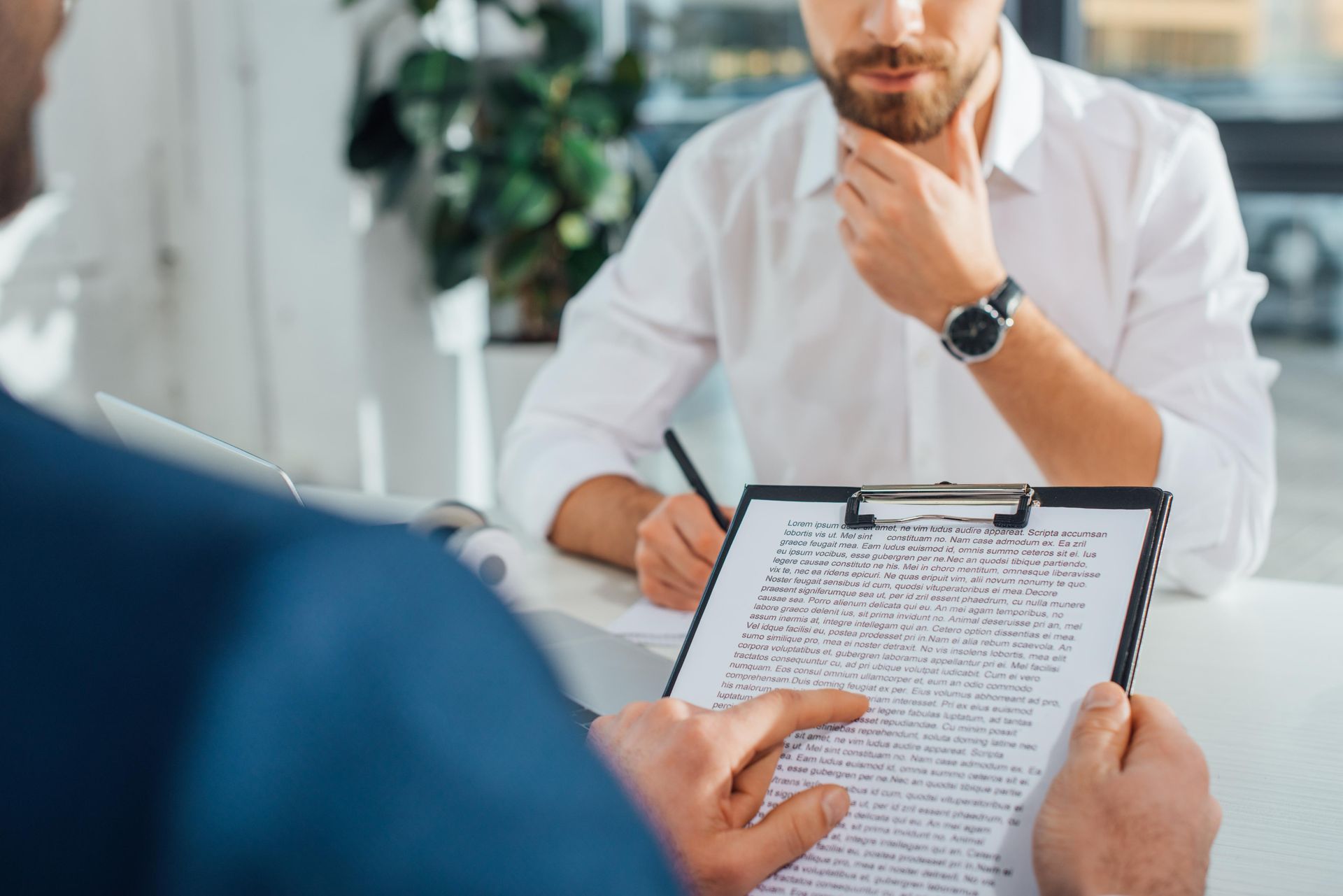 A man is sitting at a table holding a clipboard and talking to another man.