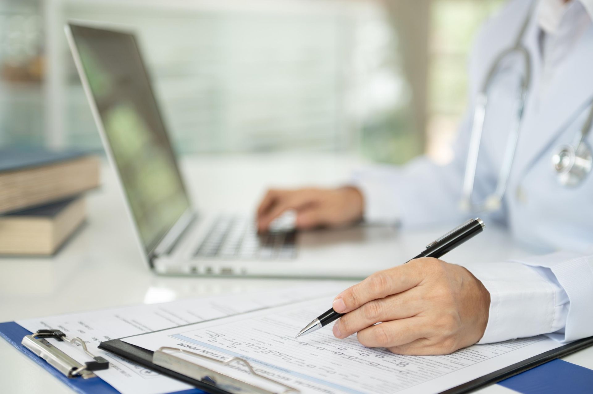 A doctor is writing on a clipboard in front of a laptop computer.