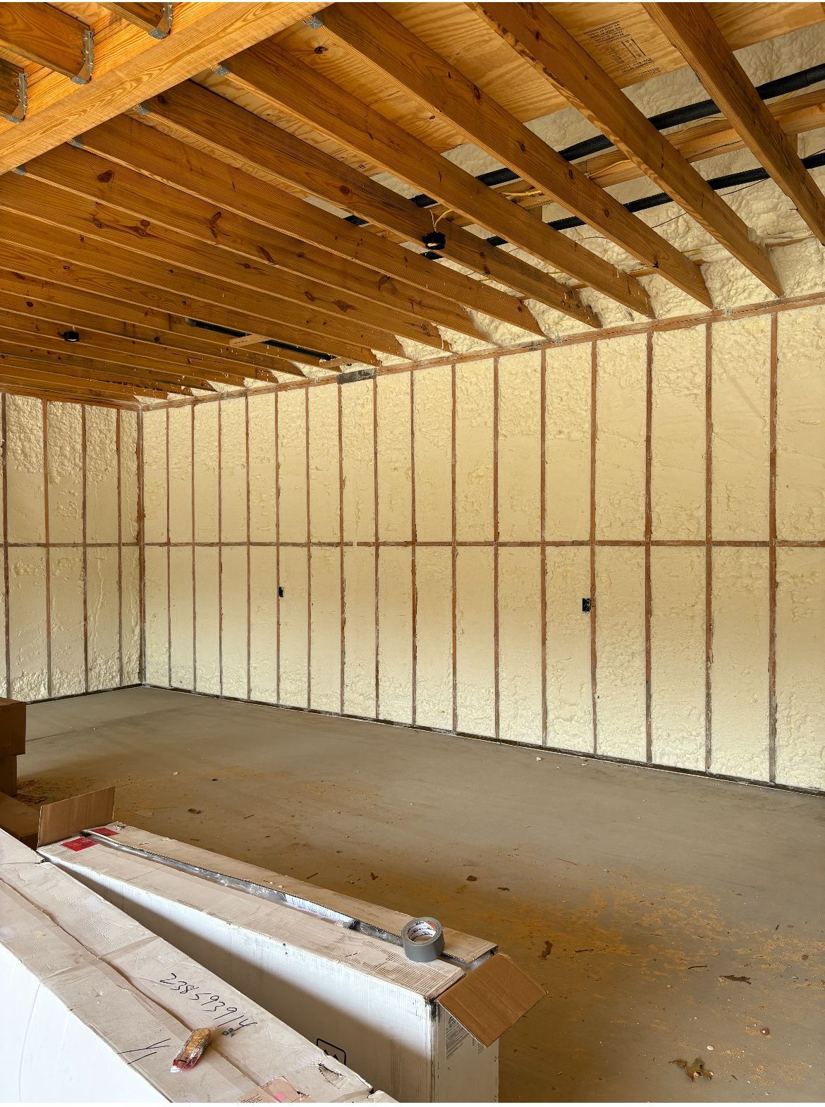 Interior view of a room with exposed wooden beams and spray foam insulation. Concrete floor and cardboard boxes.