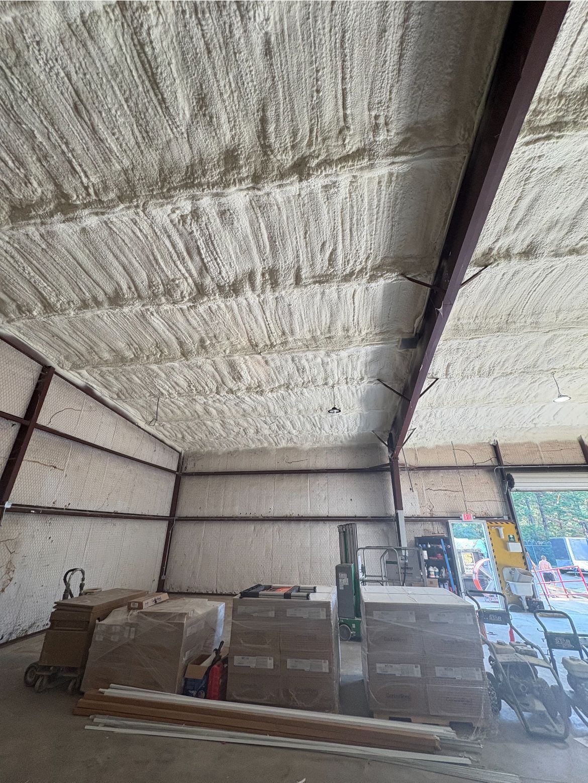 Interior of a metal building with spray foam insulation on the ceiling and walls. Construction materials are visible.