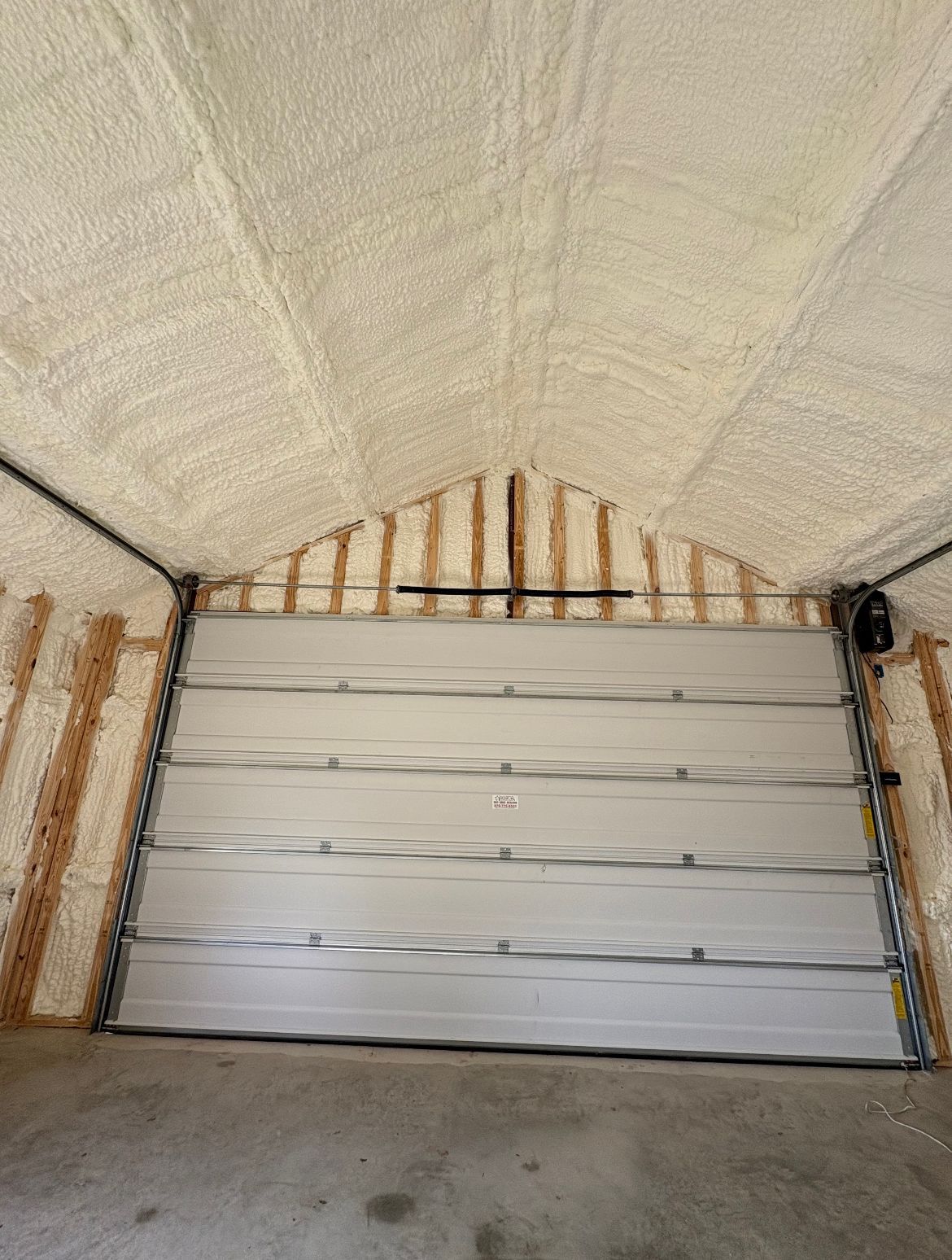 Interior of a garage with a closed white garage door. Ceiling and walls are insulated with tan foam.