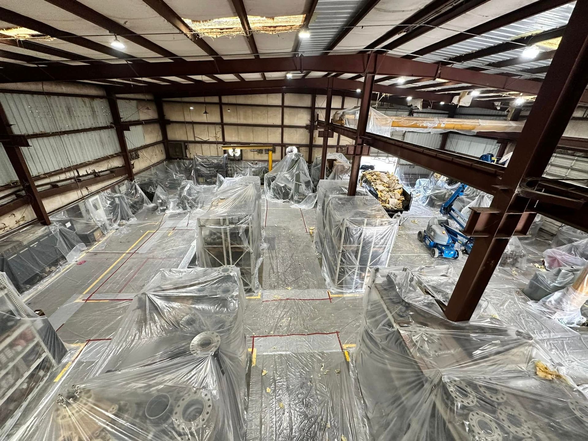 Large warehouse filled with machinery wrapped in plastic, viewed from above. Steel beams and overhead lighting present.
