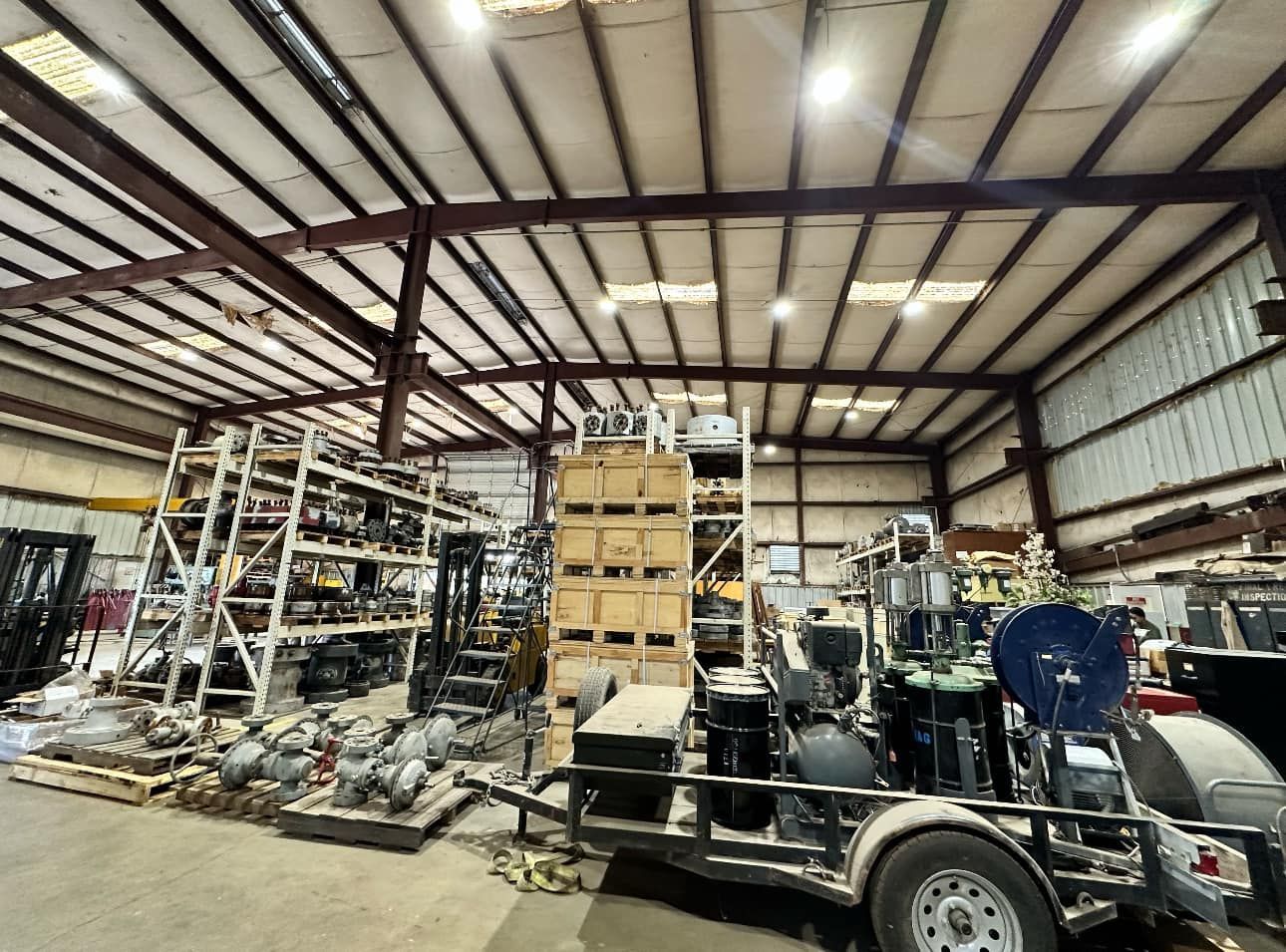 Warehouse interior filled with equipment, machinery, and shelves; a trailer with a pump is in the foreground.