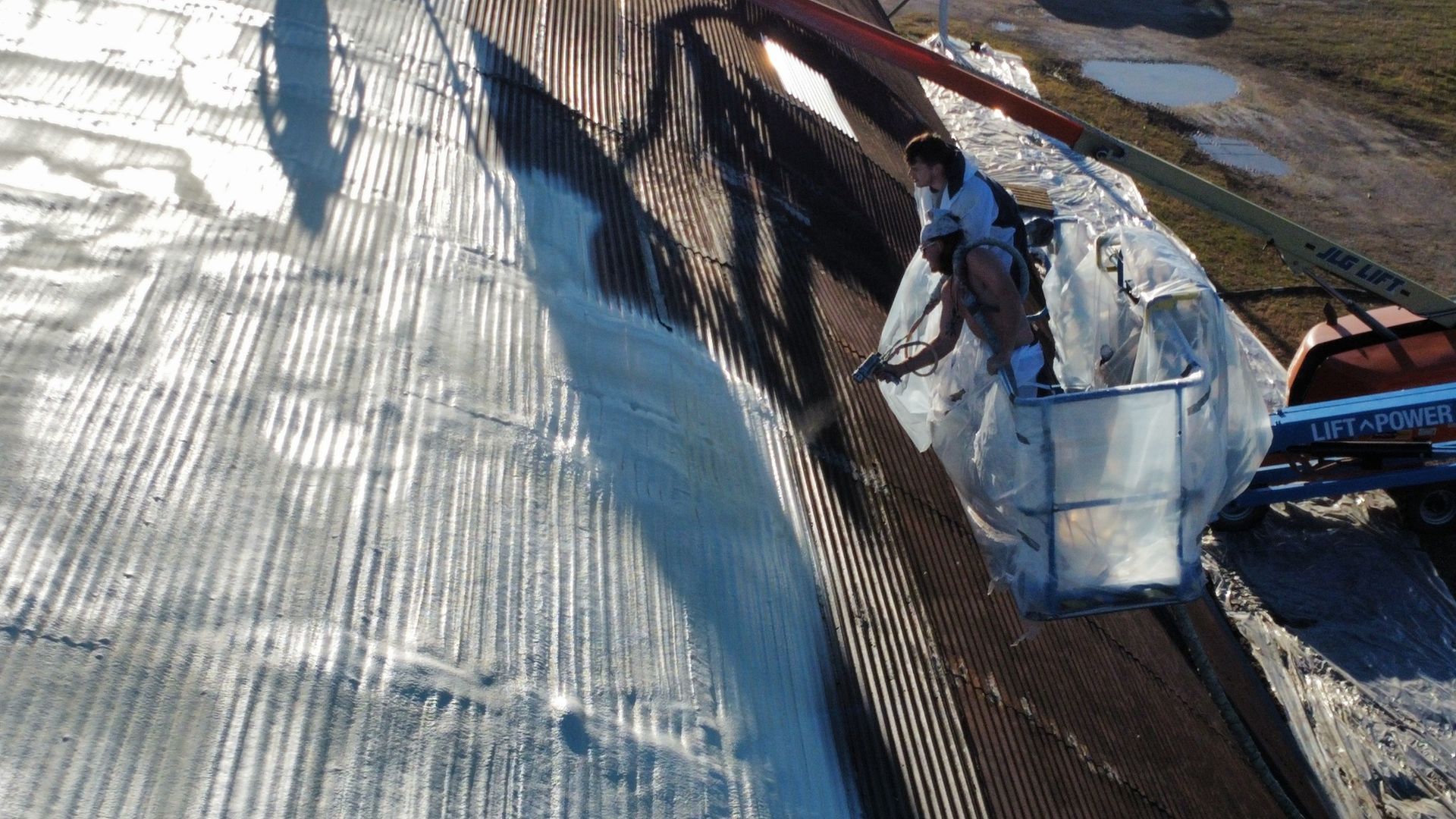 Person in lift bucket on a corrugated metal roof, applying white coating.