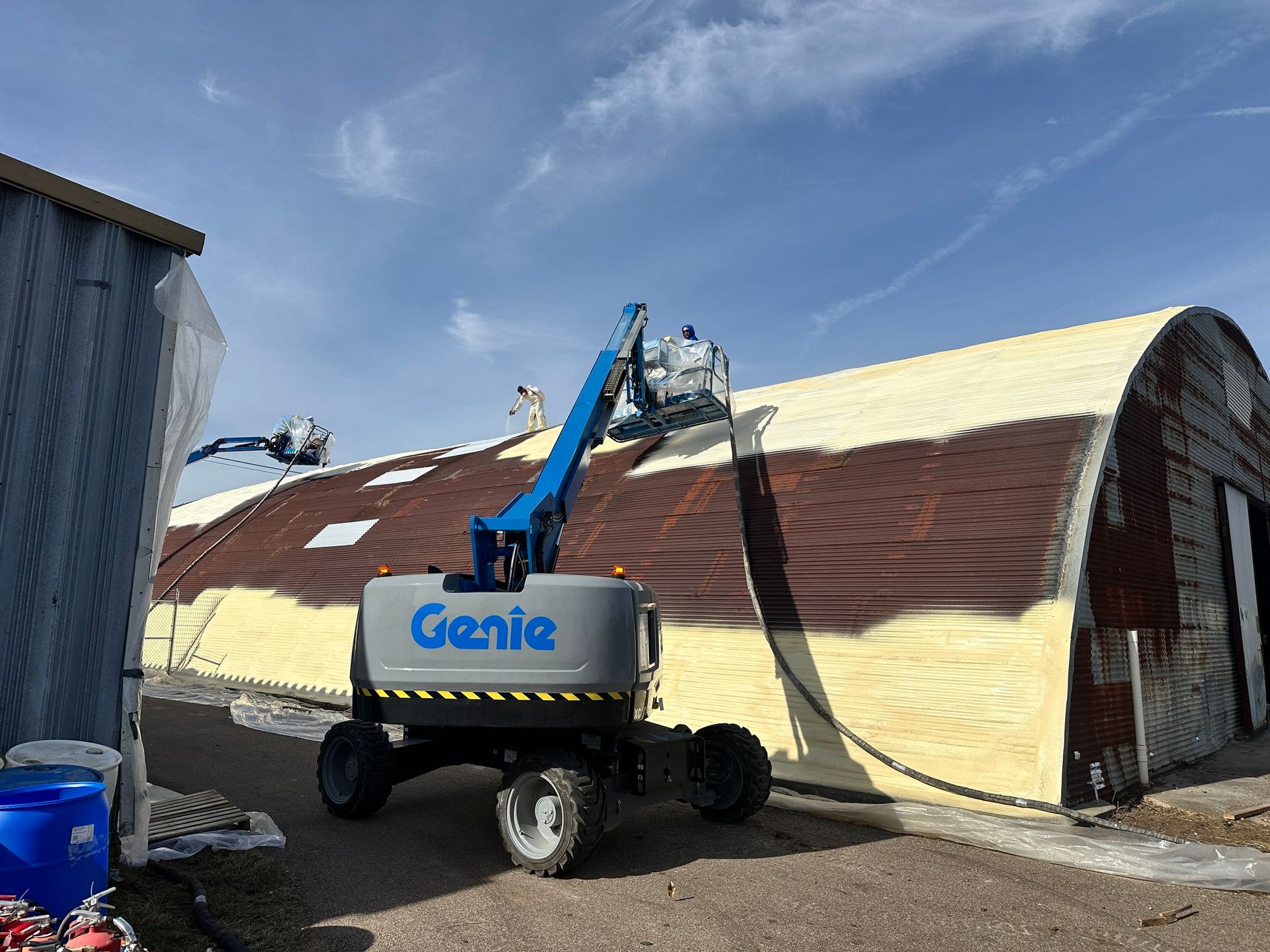 Workers spraying foam insulation on a curved metal building using a Genie lift on a sunny day.