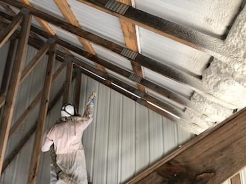Person spraying white foam insulation on the interior of a metal roof structure.
