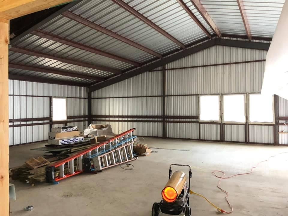 Large, empty metal building interior; construction in progress. Ladder, materials, and space heater visible.