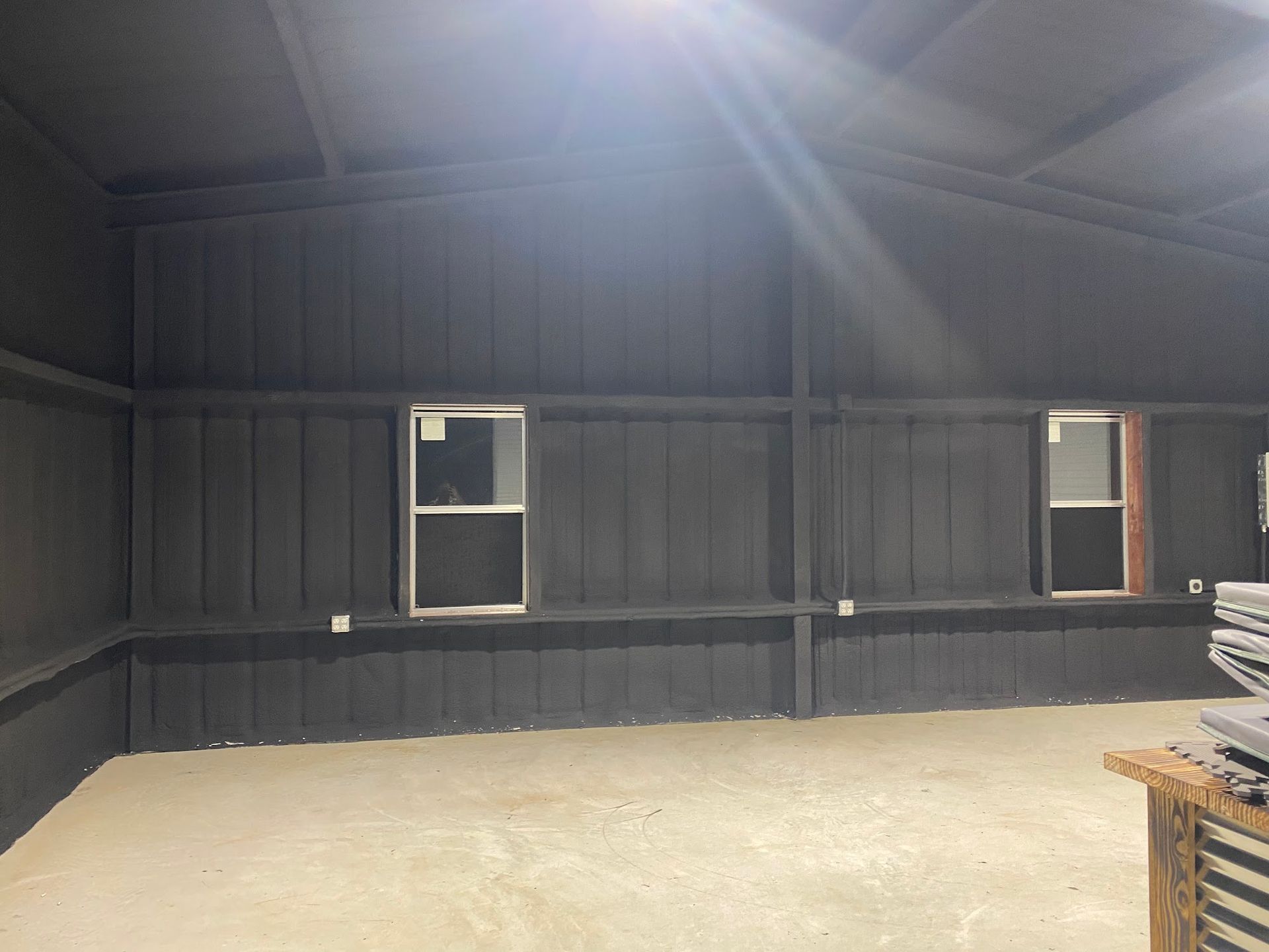 Interior of a dark-painted building with two windows. The room has a concrete floor and a wood shelf.