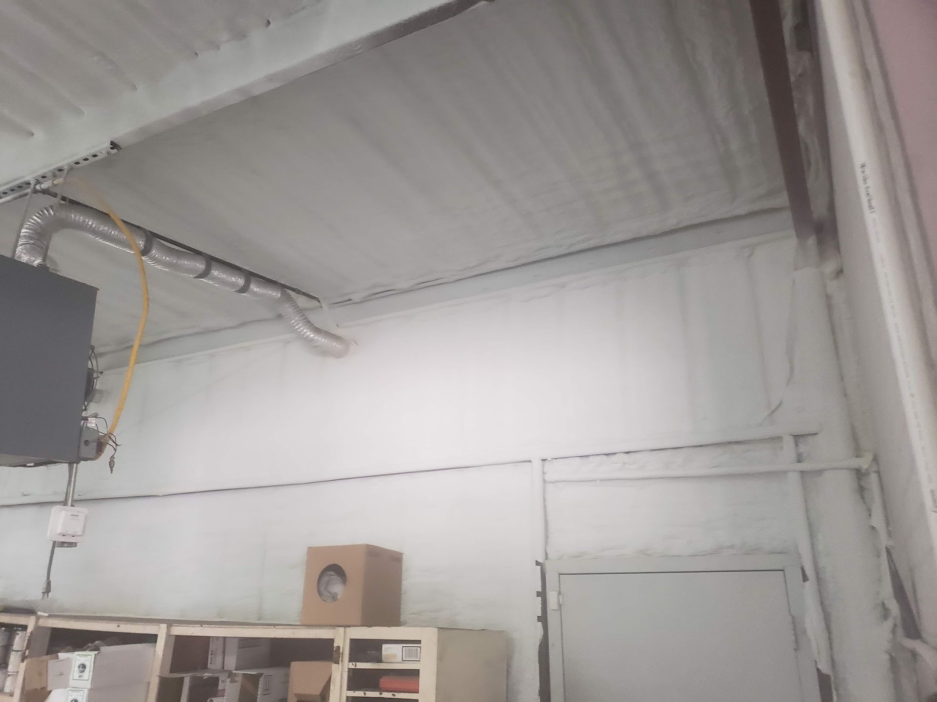 White foam insulation covers the ceiling and walls of a commercial space. Brown cardboard box on a shelf.