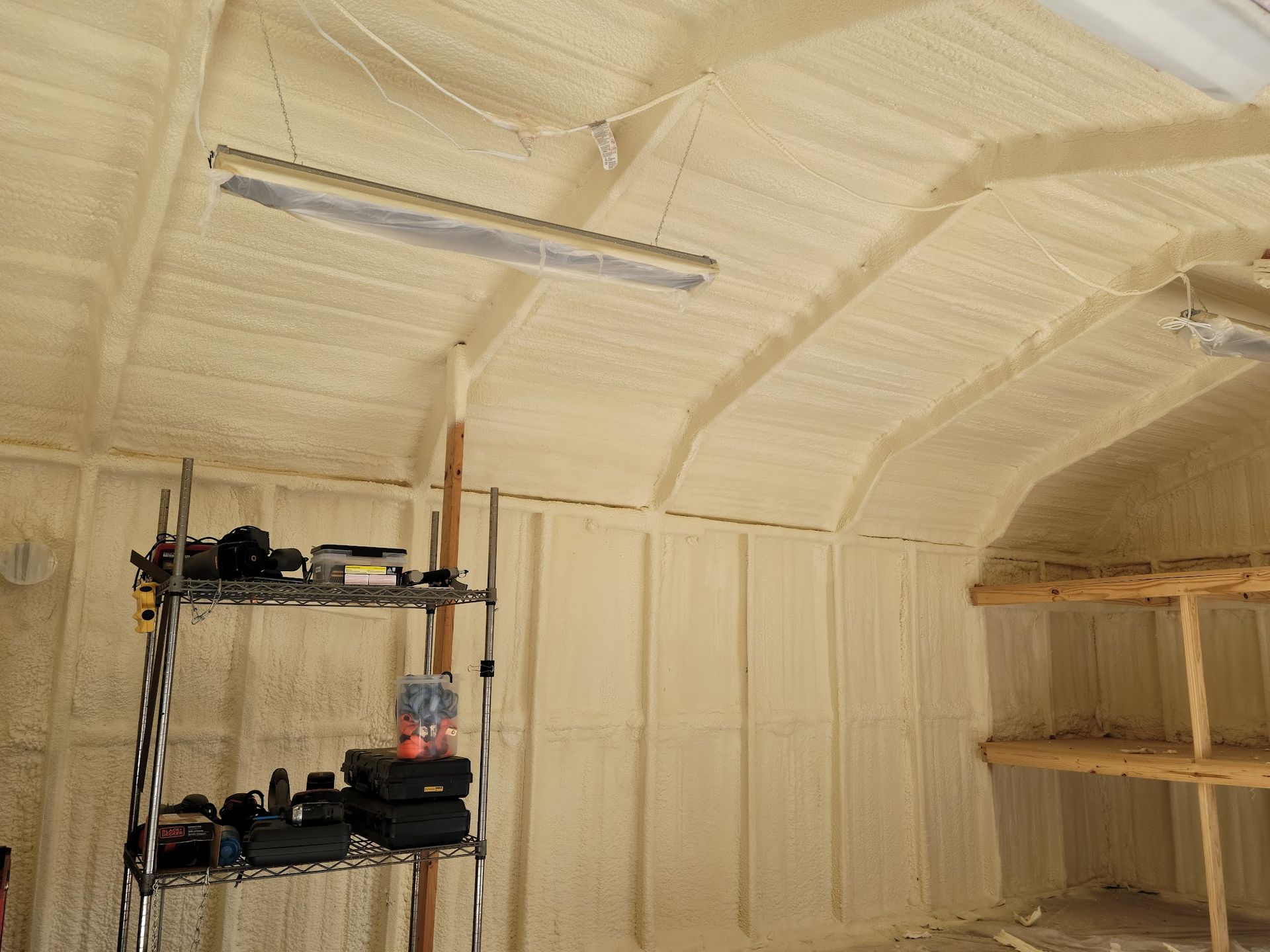 Garage interior with tan spray foam insulation on walls and arched ceiling; shelving units and work lights are visible.