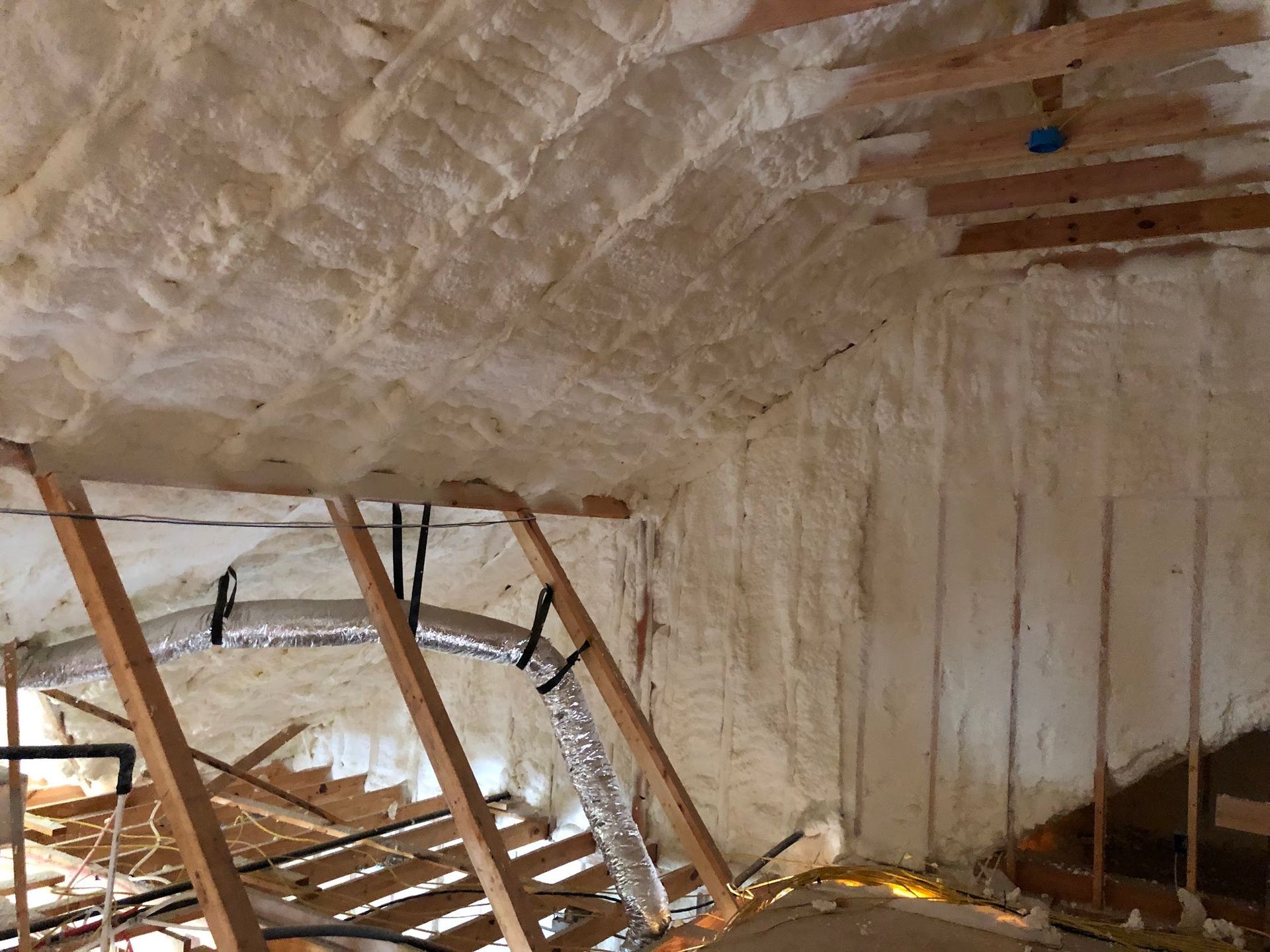 Attic interior with spray foam insulation on ceiling and walls. Wooden rafters and ductwork visible.