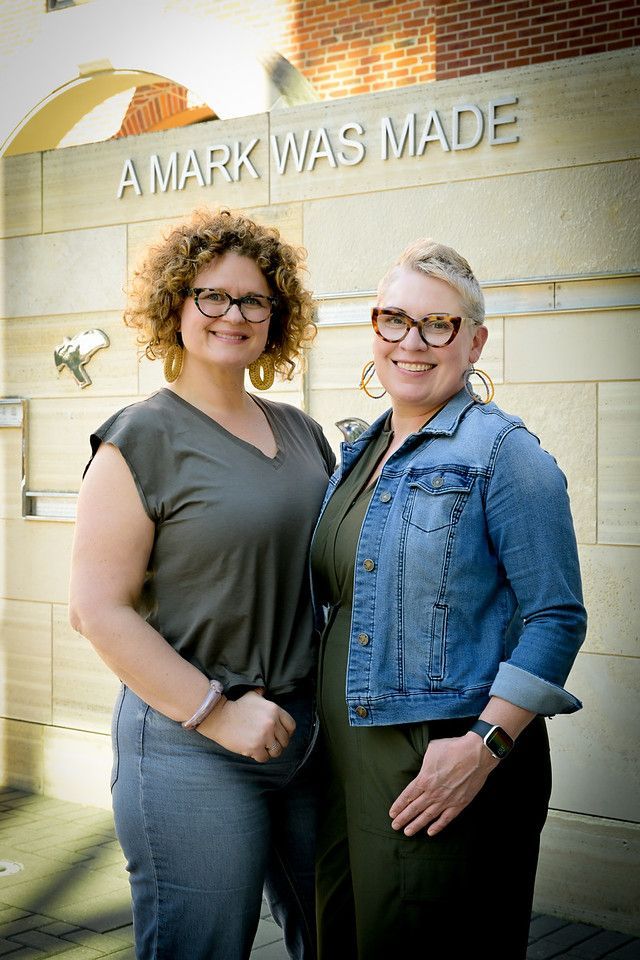 Two women are standing next to each other in front of a sign that says a mark was made.