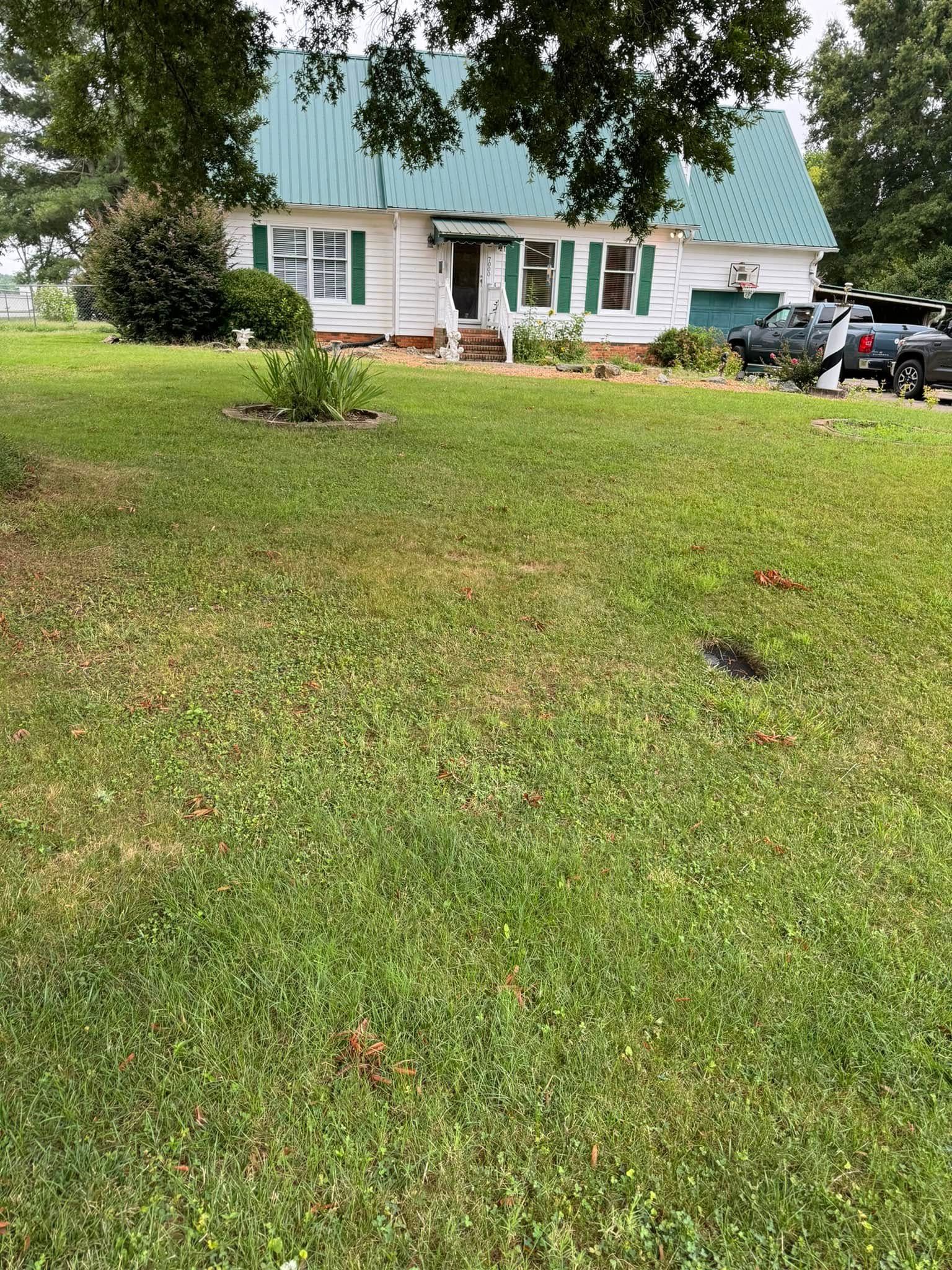 A white house with green shutters and a green roof sits on a grassy lawn. A few vehicles are parked in the background.