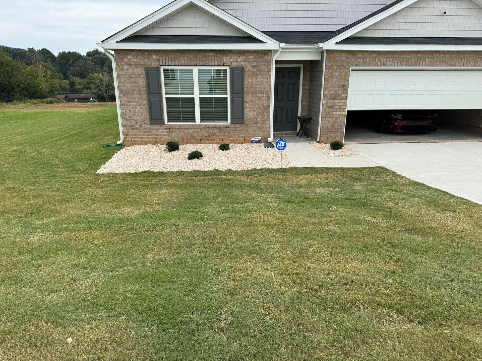 Brick house with gray roof, garage, and lawn. Front yard features a gravel bed with small green plants.