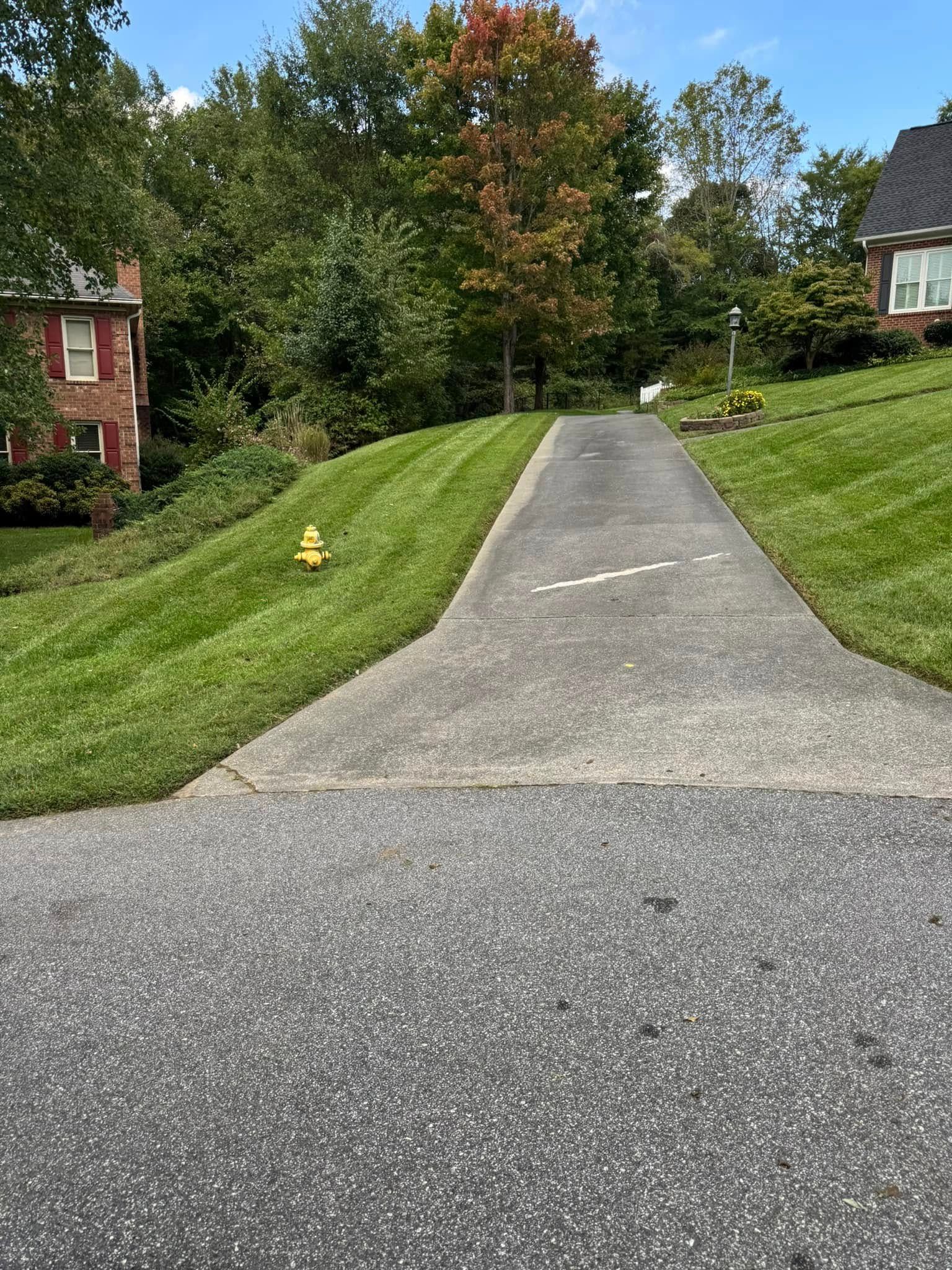 Concrete driveway leading uphill, flanked by green grass. A brick building and trees are visible in the background.