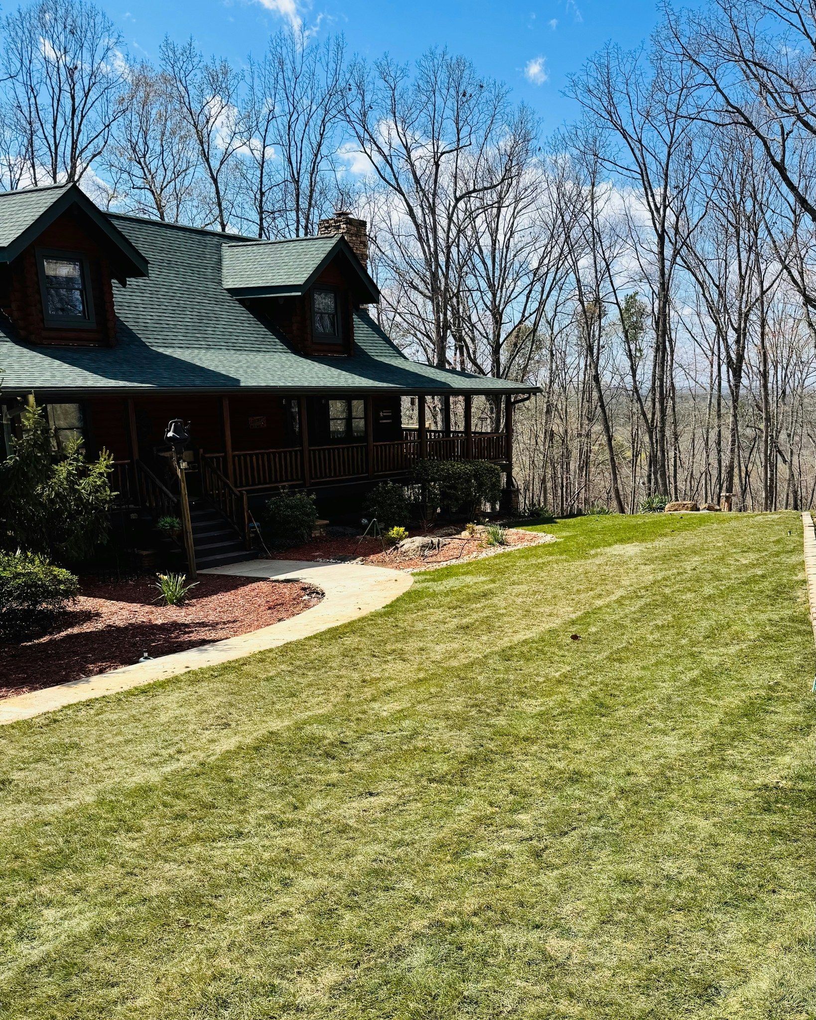 Cozy cabin with green roof and wraparound porch sits on a grassy hill. Trees line the background under a bright blue sky.