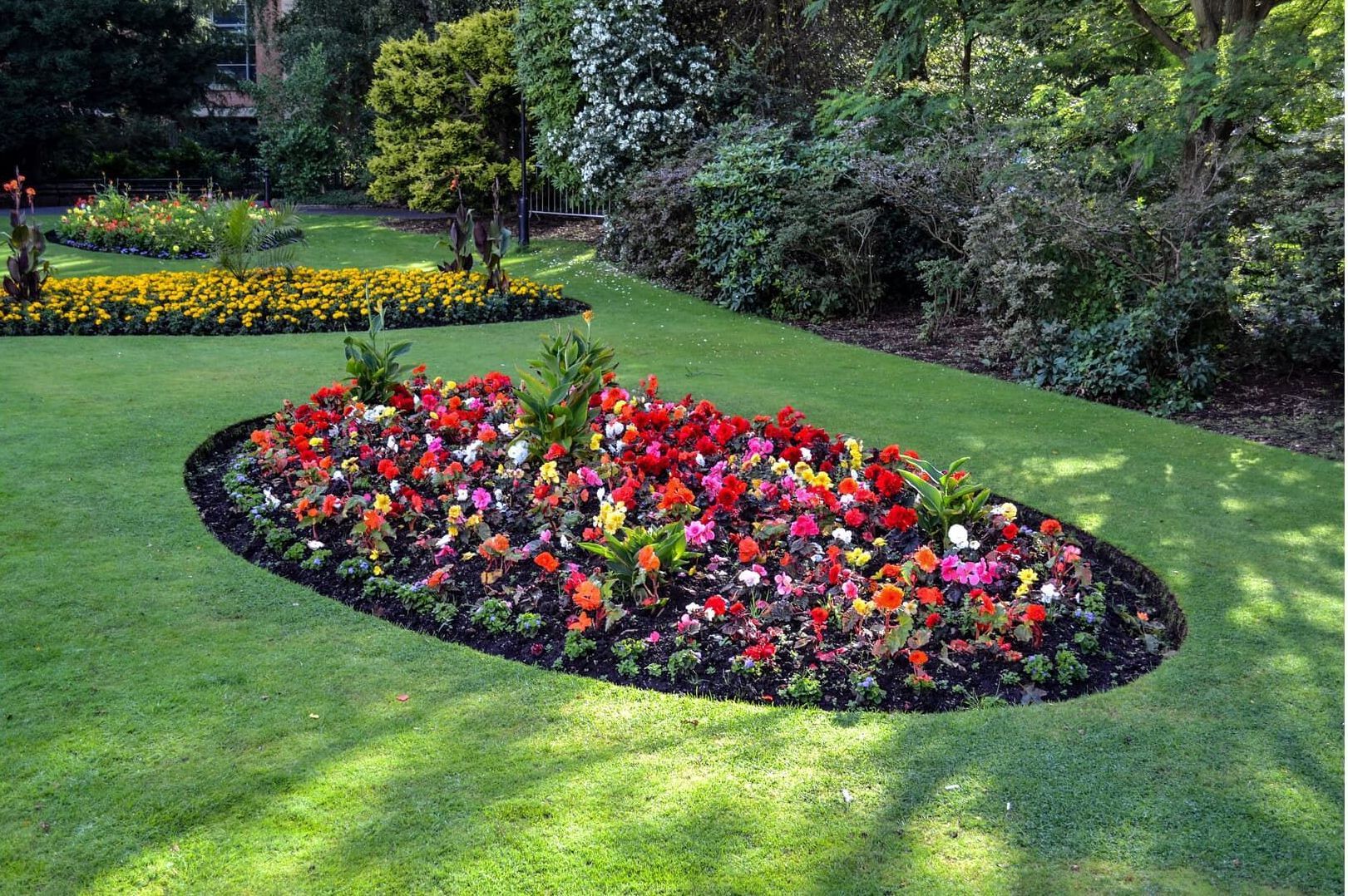 Oval flower bed overflowing with colorful flowers, set in green lawn. More flower beds and trees in the background.