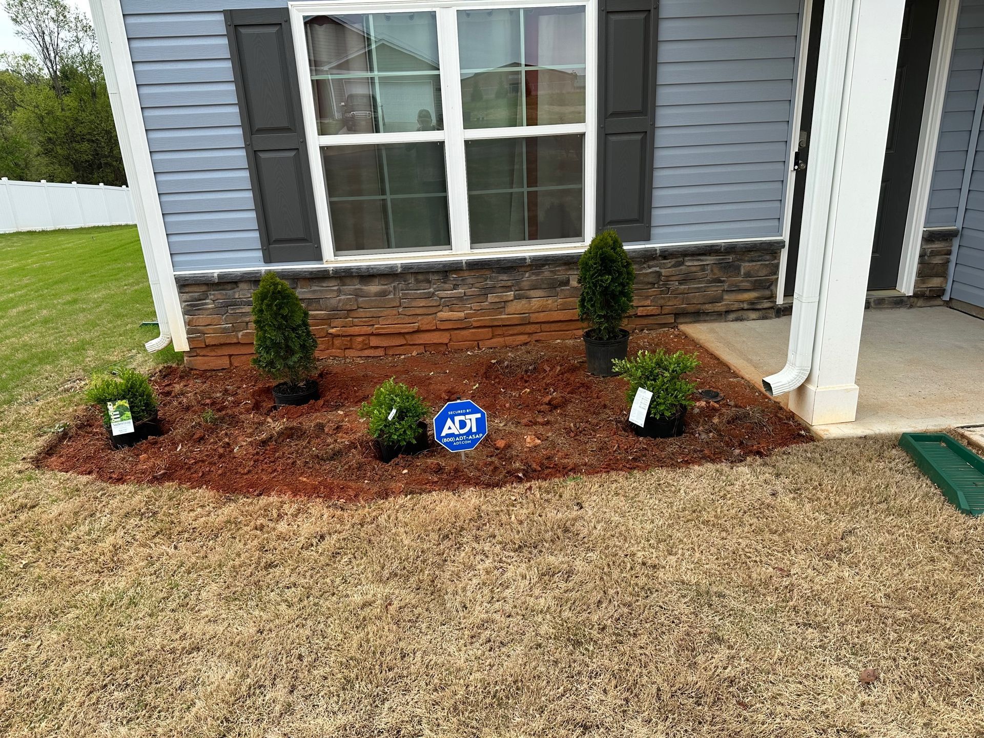 A house exterior with a small garden bed of new plants and wood chips; blue siding, a large window, and an ADT security sign.
