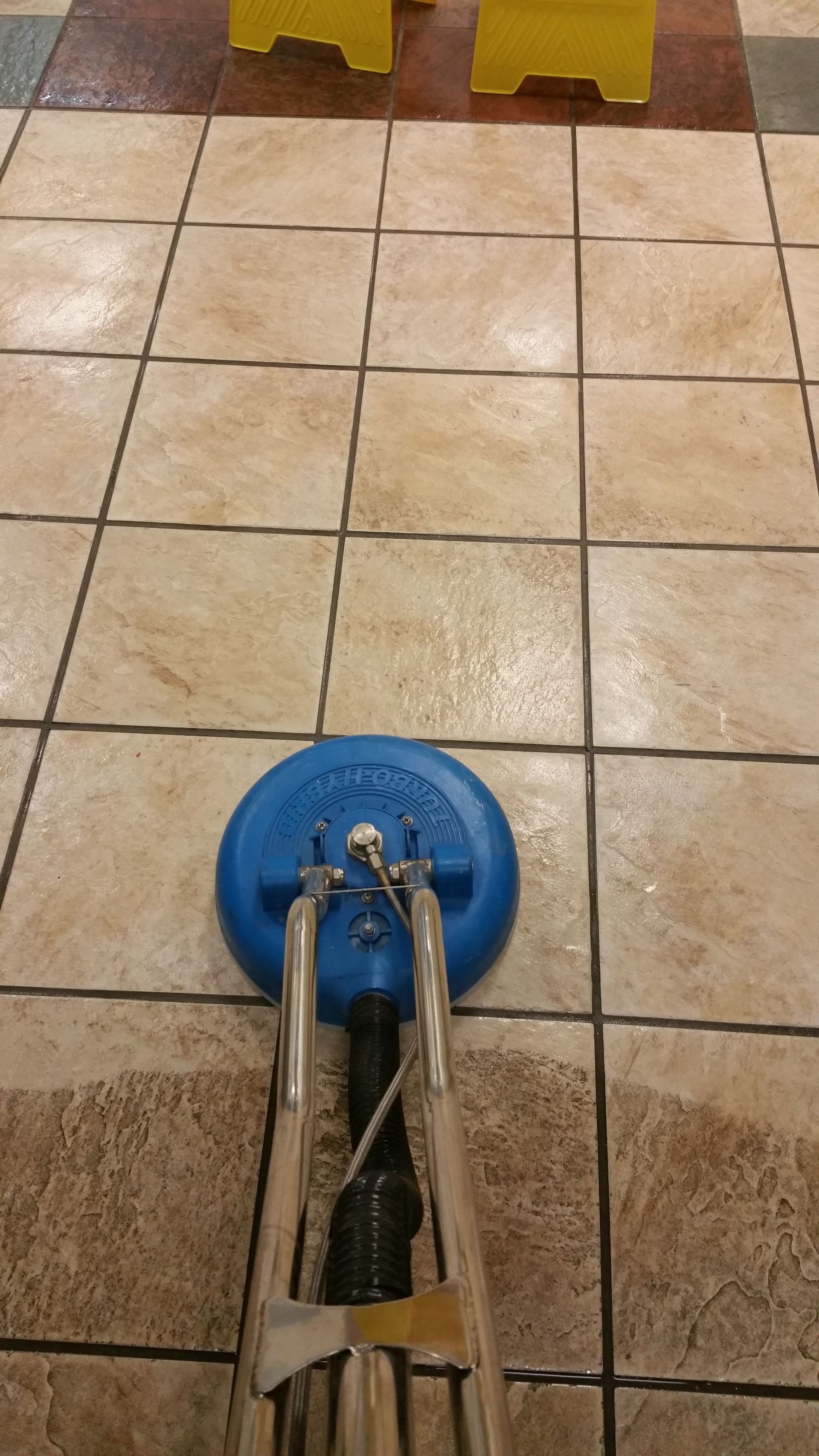 Blue tile-cleaning machine on a wet tiled floor with yellow barriers in the background
