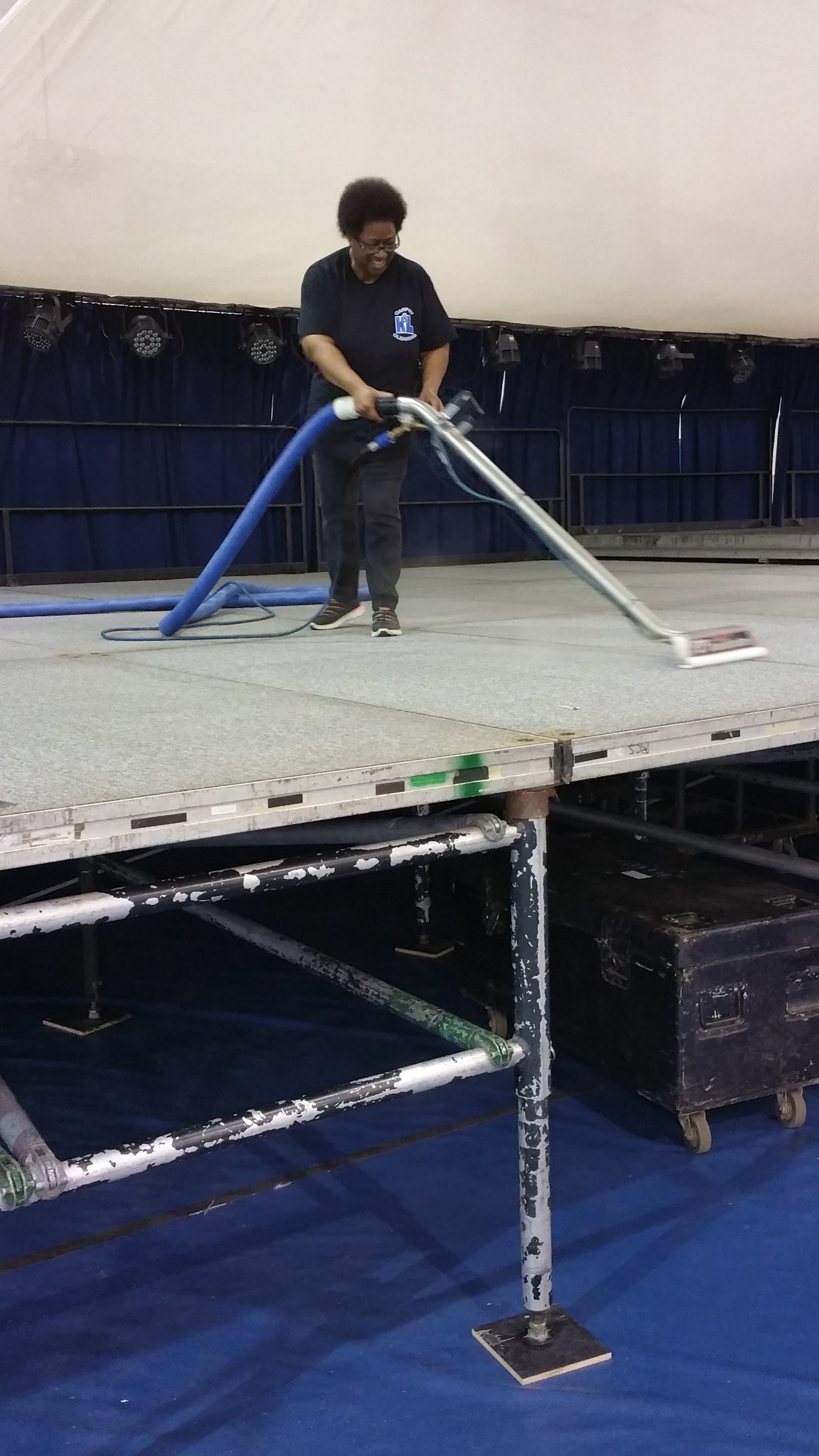 Person on a stage vacuuming a gray floor with a blue hose and silver wand