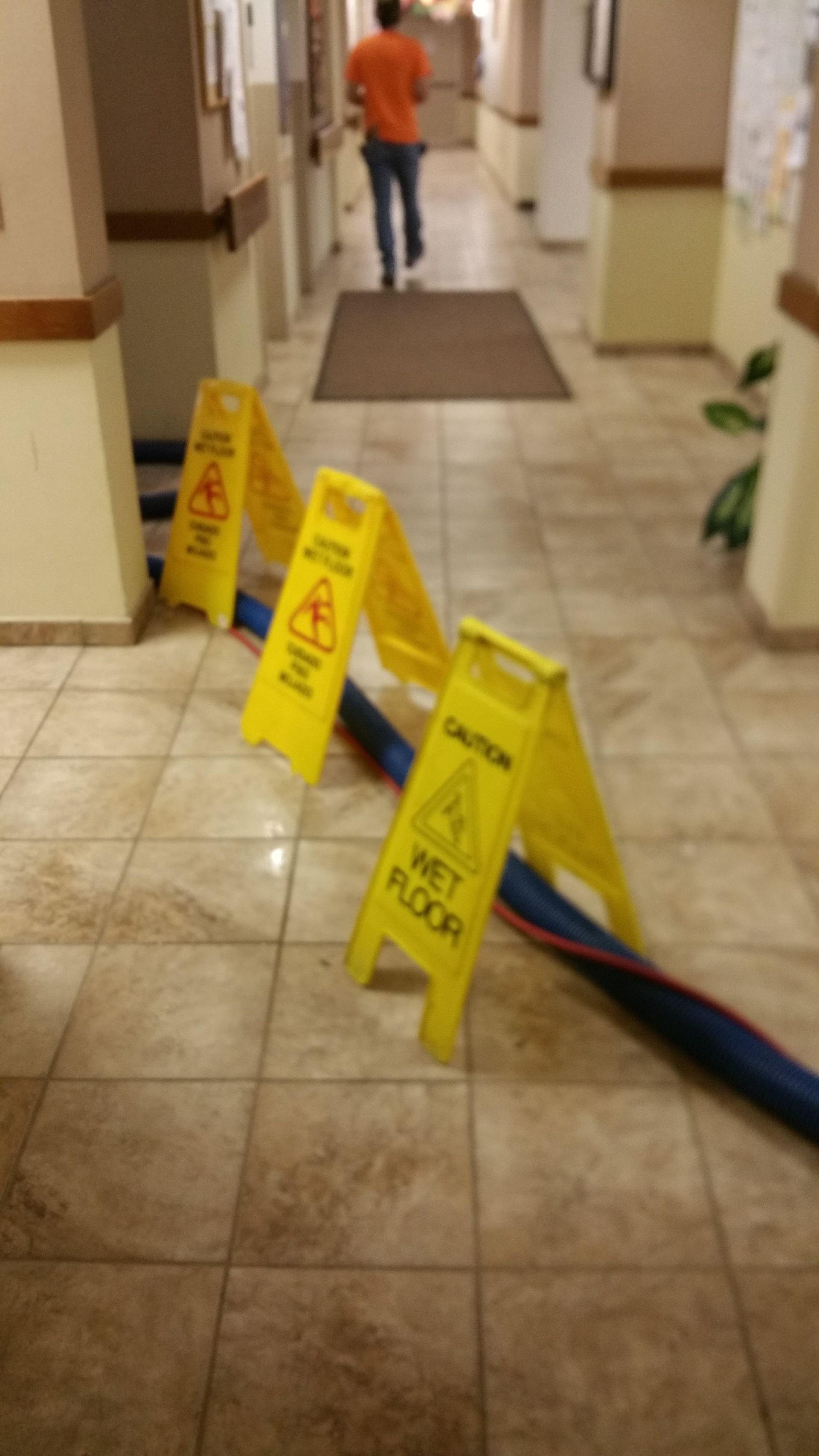 Yellow wet-floor signs in a hallway, with a hose across the tiled floor and a person walking away.