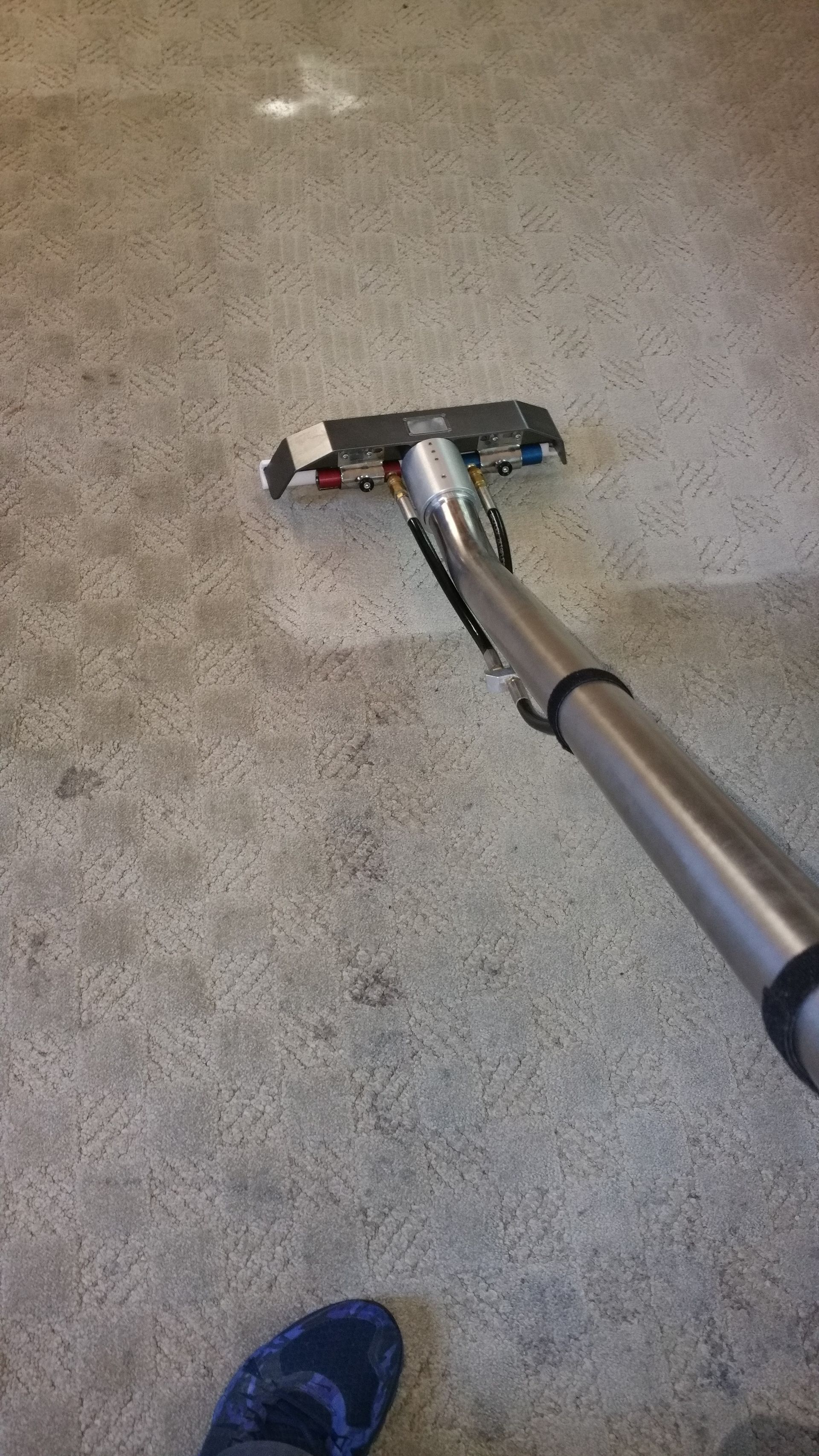 Carpet being cleaned with a silver vacuum nozzle; a blue shoe is visible at the bottom left.