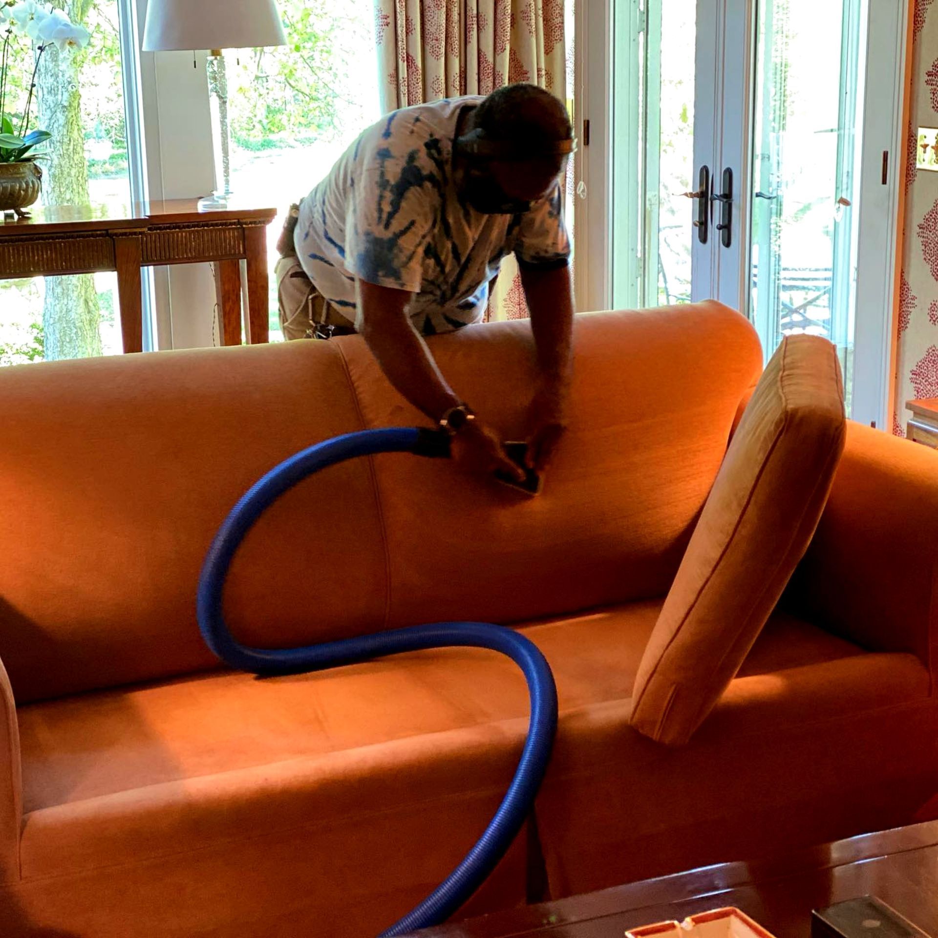 A person cleans an orange couch with a blue vacuum hose in a living room.