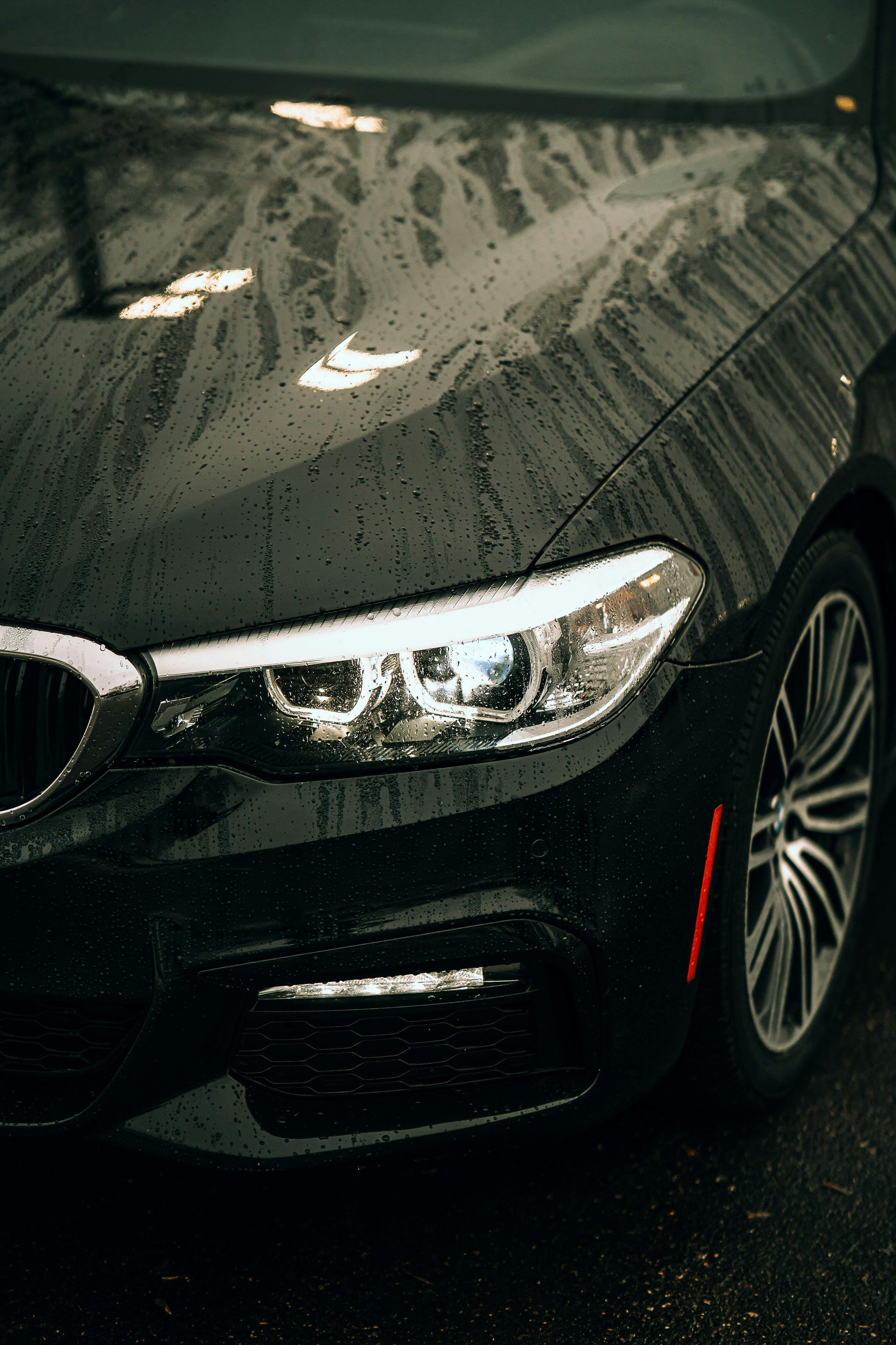 Black BMW car, wet with raindrops, close-up of headlight and wheel.