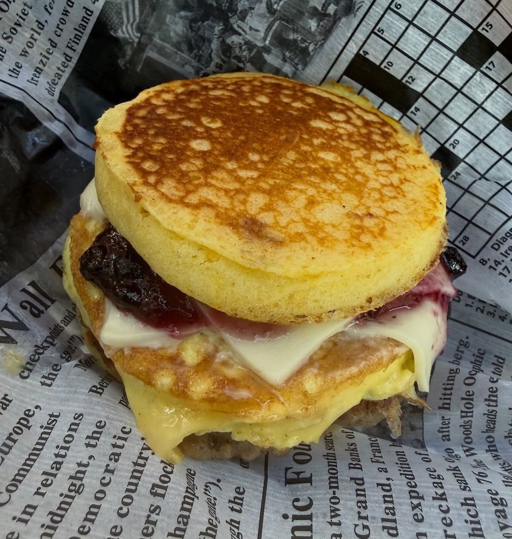 Stacked breakfast sandwich with toasted muffin, egg, cheese, and bacon on newspaper wrap