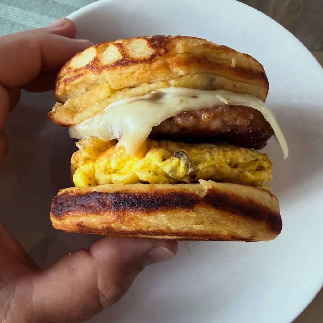 Hand holding a stacked breakfast sandwich with egg, sausage, and melted cheese on a white plate