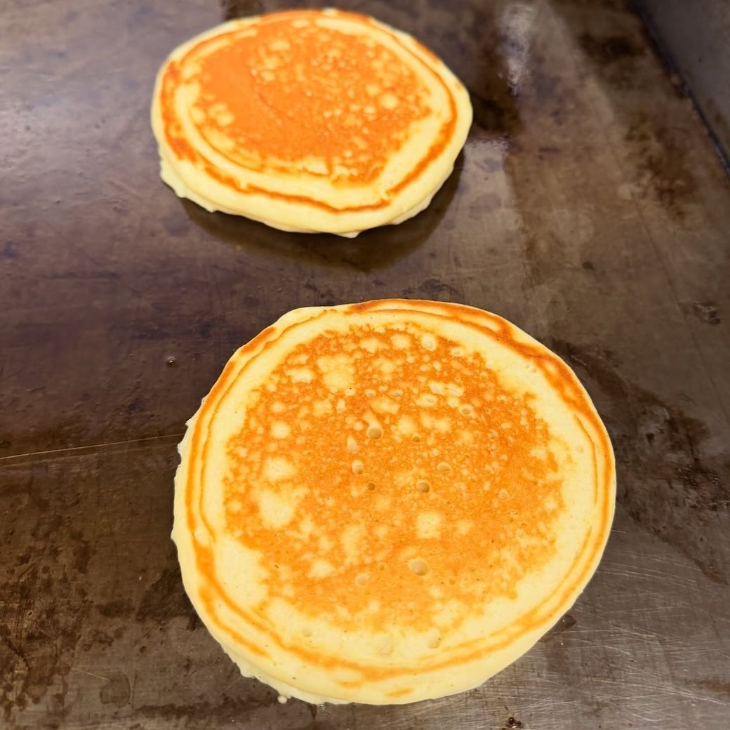 Two pancakes cooking on a griddle, one slightly browned more than the other