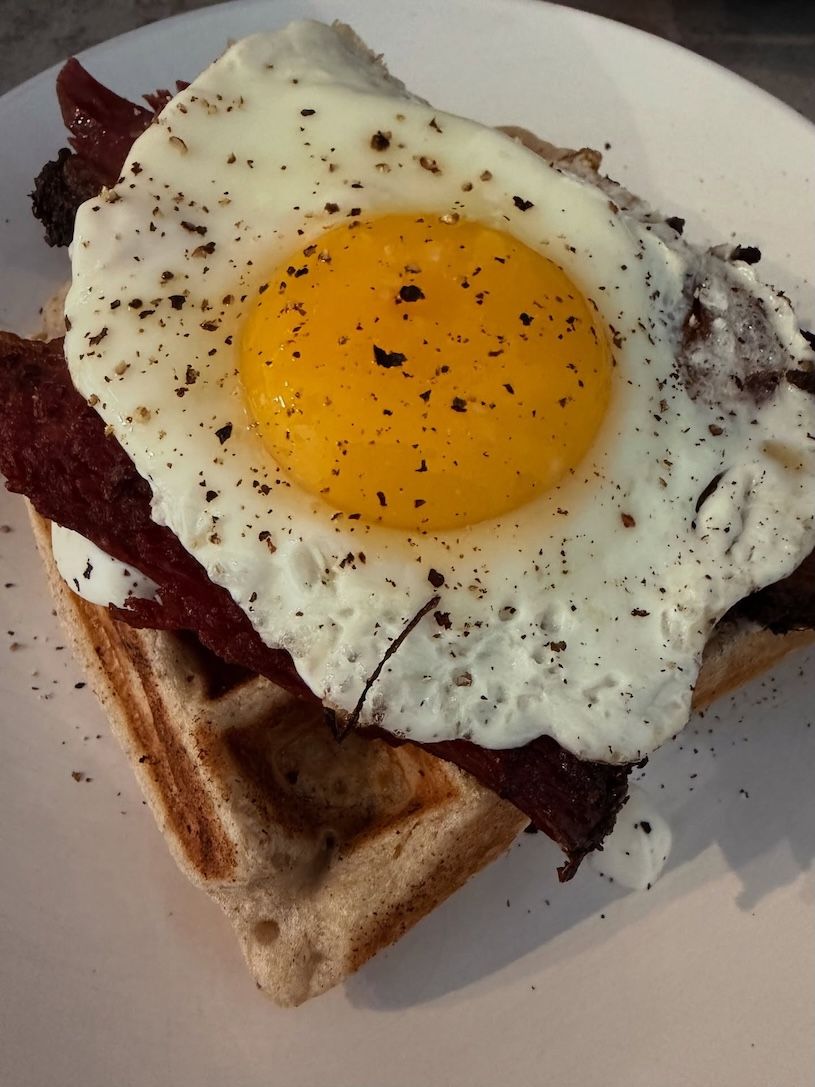 Belgian waffle topped with a sunny-side-up egg, bacon, and pepper on a white plate
