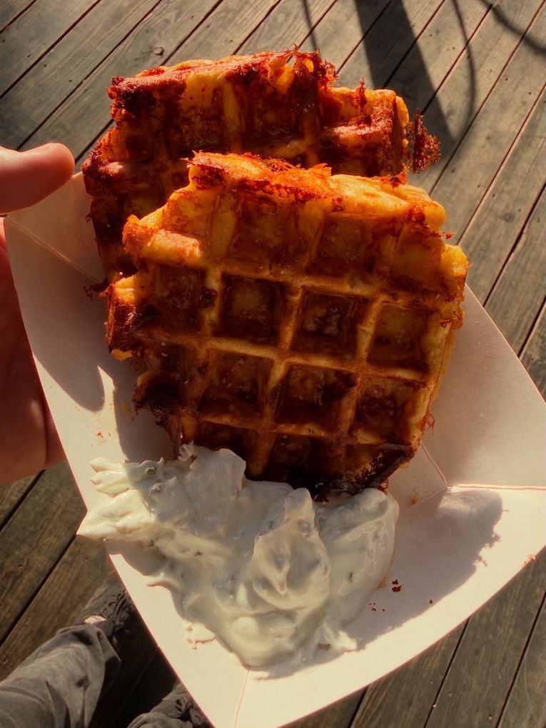 Two waffles on a paper plate, one topped with syrup, held outdoors on a wooden deck.