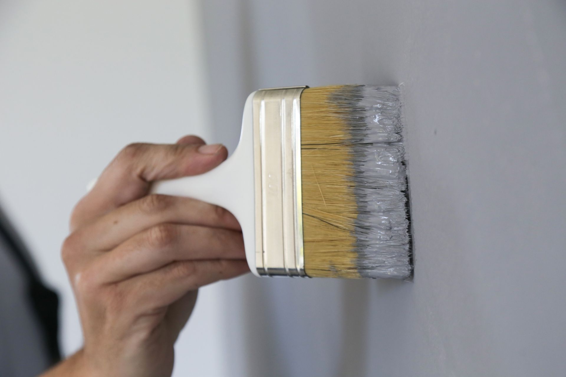 Hand using a paintbrush to apply paint on a light gray wall. Hand using a paintbrush to apply paint on a light gray wall.