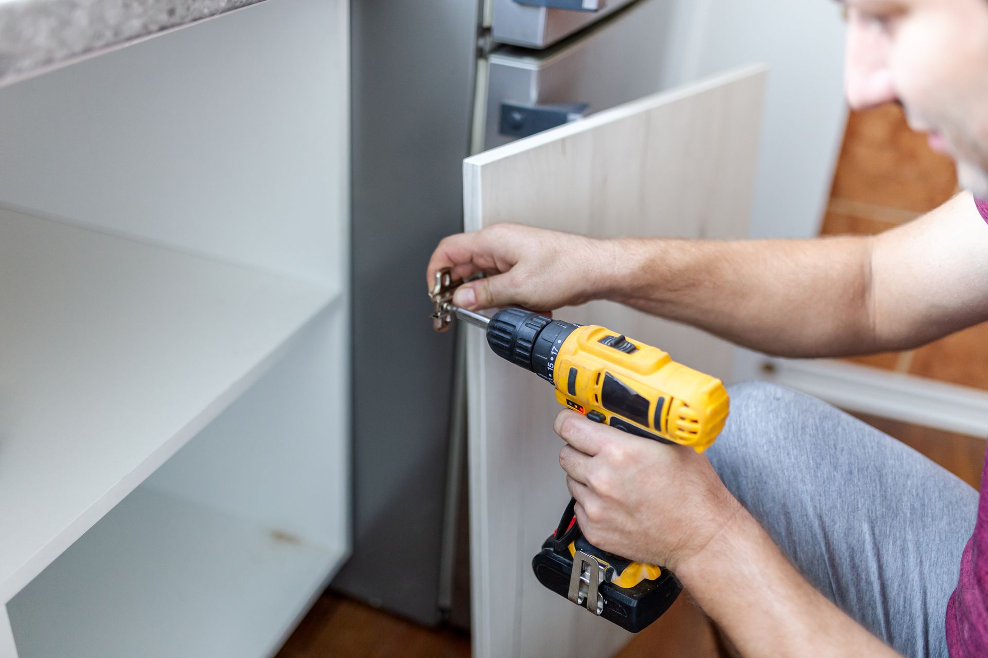 Person using a yellow drill to attach a cabinet door hinge in a white kitchen cabinet.