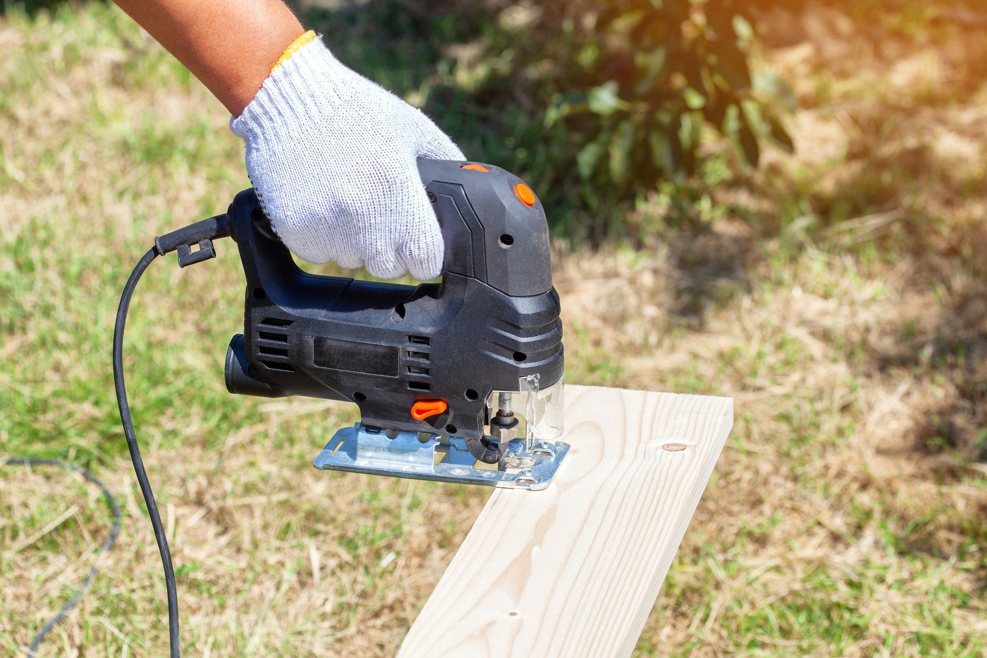 Hand with glove using a black jigsaw to cut a piece of light-colored wood in a grassy outdoor setting. Hand with glove using a black jigsaw to cut a piece of light-colored wood in a grassy outdoor setting.
