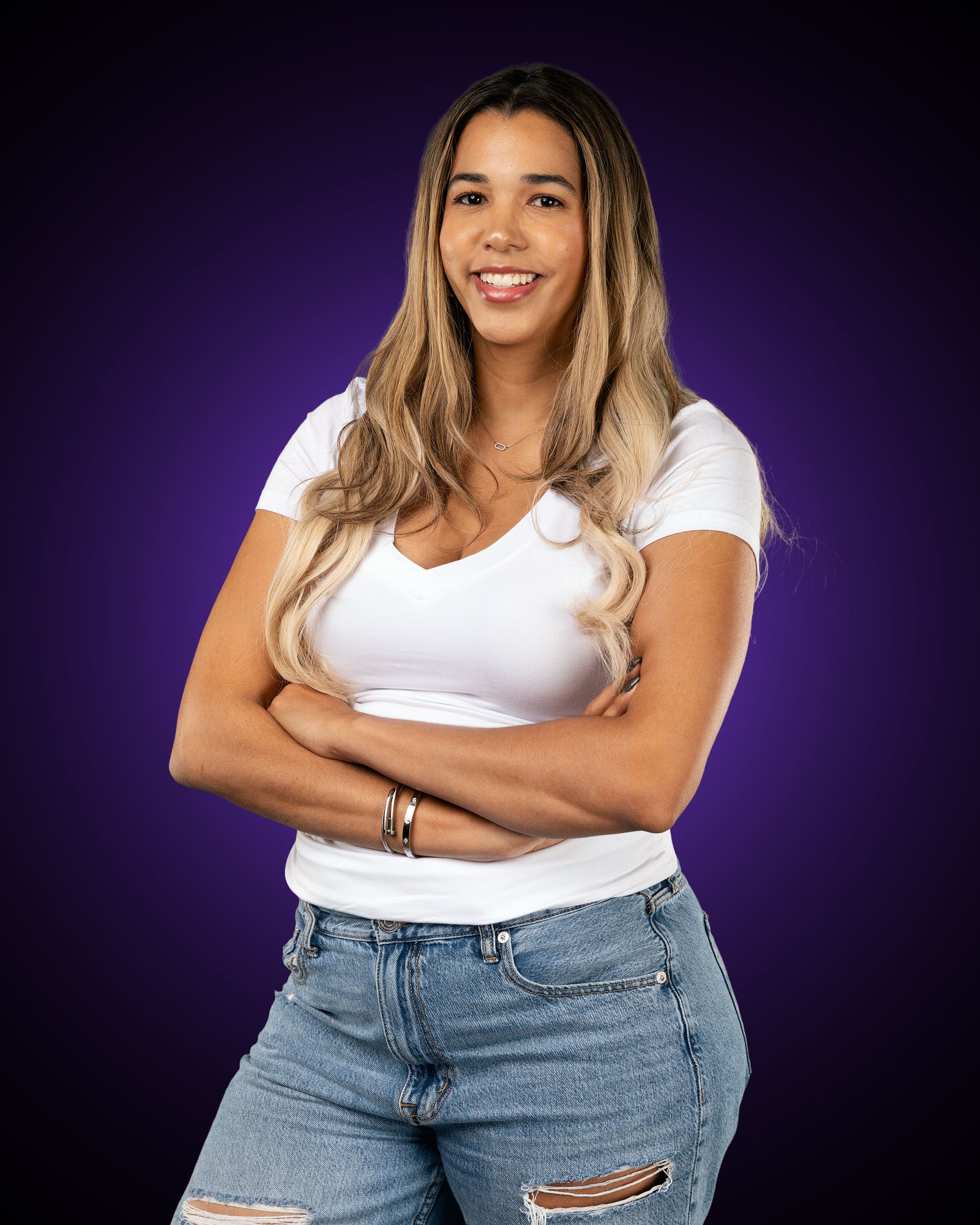 Woman in purple Sharks Cheer jacket smiles, arms crossed on a grassy field, with a whistle around her neck.