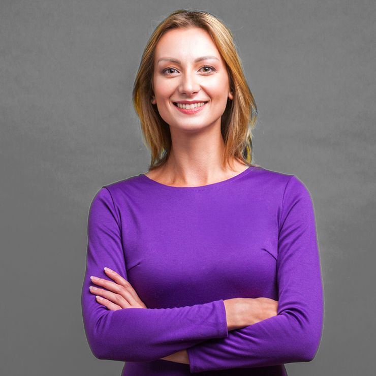 Woman with blonde hair wearing a purple long-sleeved shirt, smiling with arms crossed in front of a gray background.
