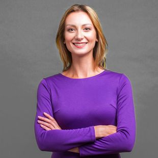 Woman with blonde hair wearing a purple long-sleeved shirt, smiling with arms crossed in front of a gray background.