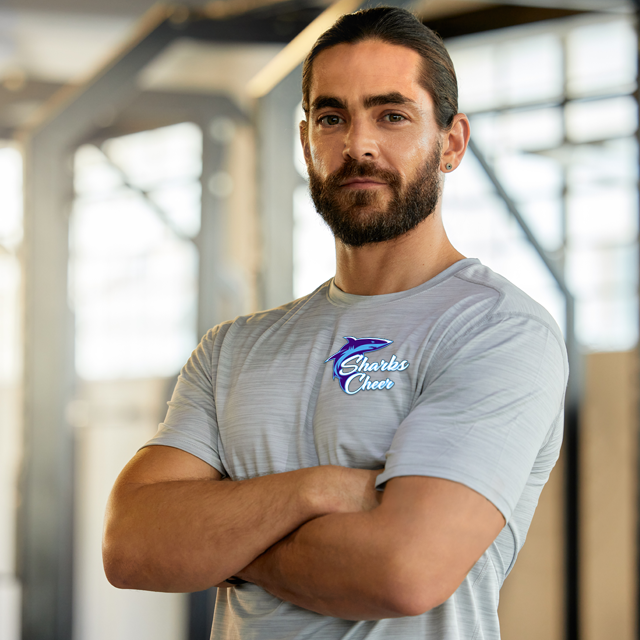 A man with a beard and long hair, wearing a gray shirt with a Sharks Cheer logo, arms crossed, in a gym.