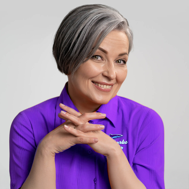 Woman with gray hair wearing a purple shirt, smiling with hands clasped. Set against a gray background.
