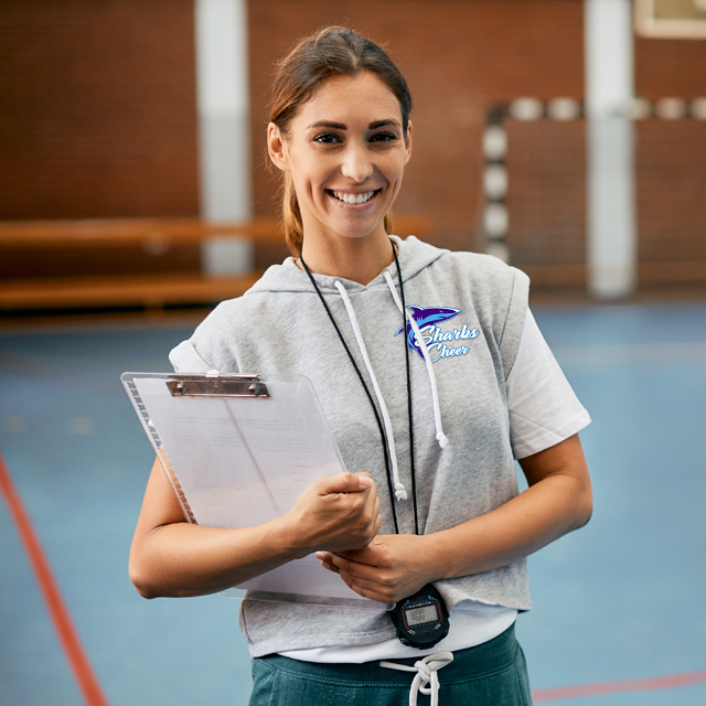 Woman coach smiling, holding a clipboard and stopwatch, in a gymnasium.