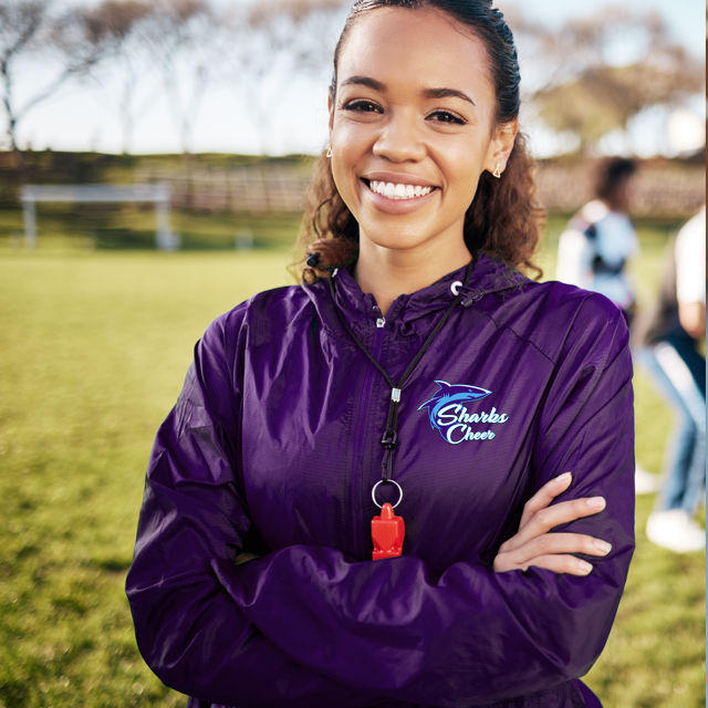 Woman in purple Sharks Cheer jacket smiles, arms crossed on a grassy field, with a whistle around her neck.